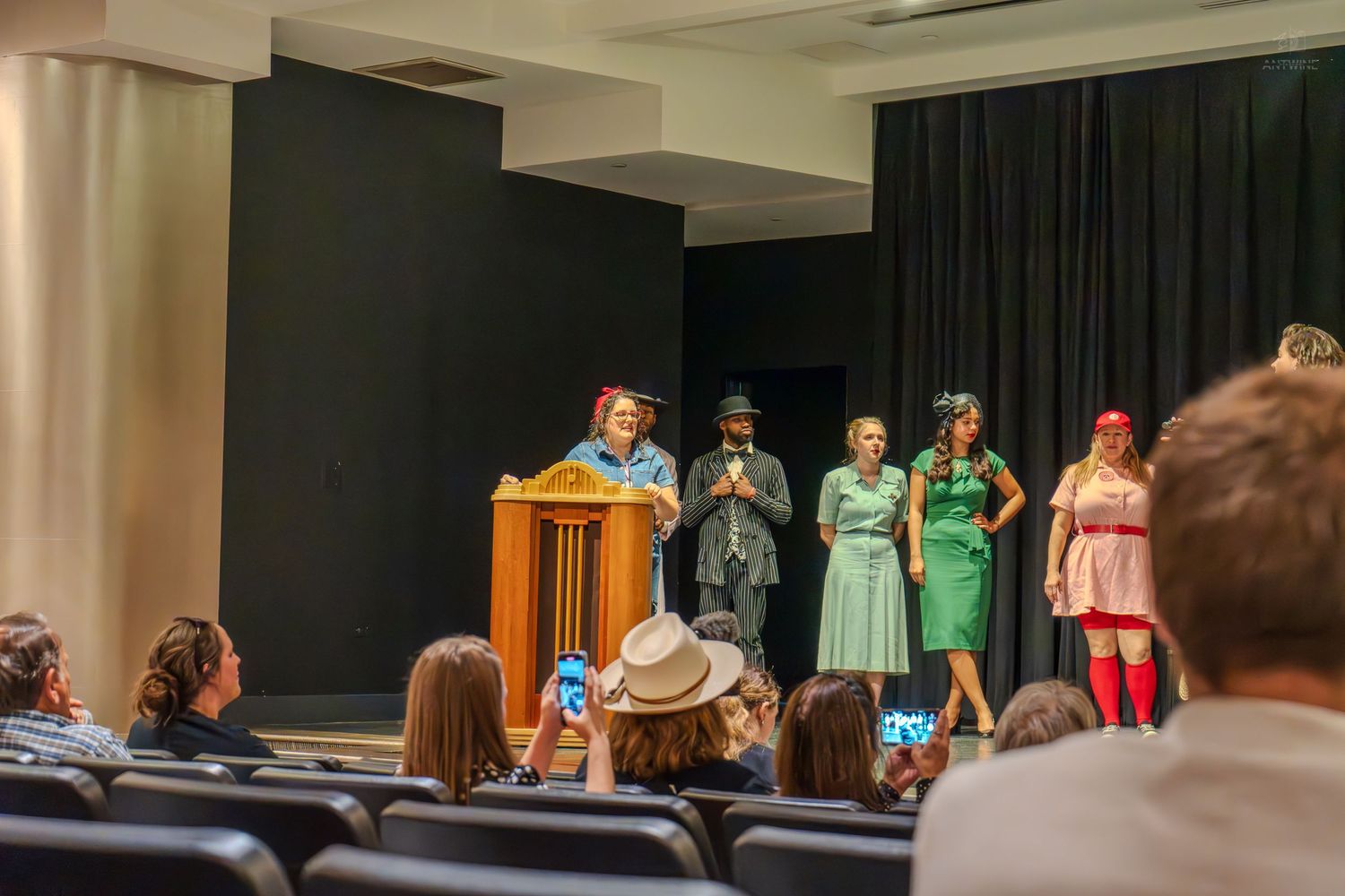 Costumed performers on stage at a theater event with audience members seated in rows watching the presentation.