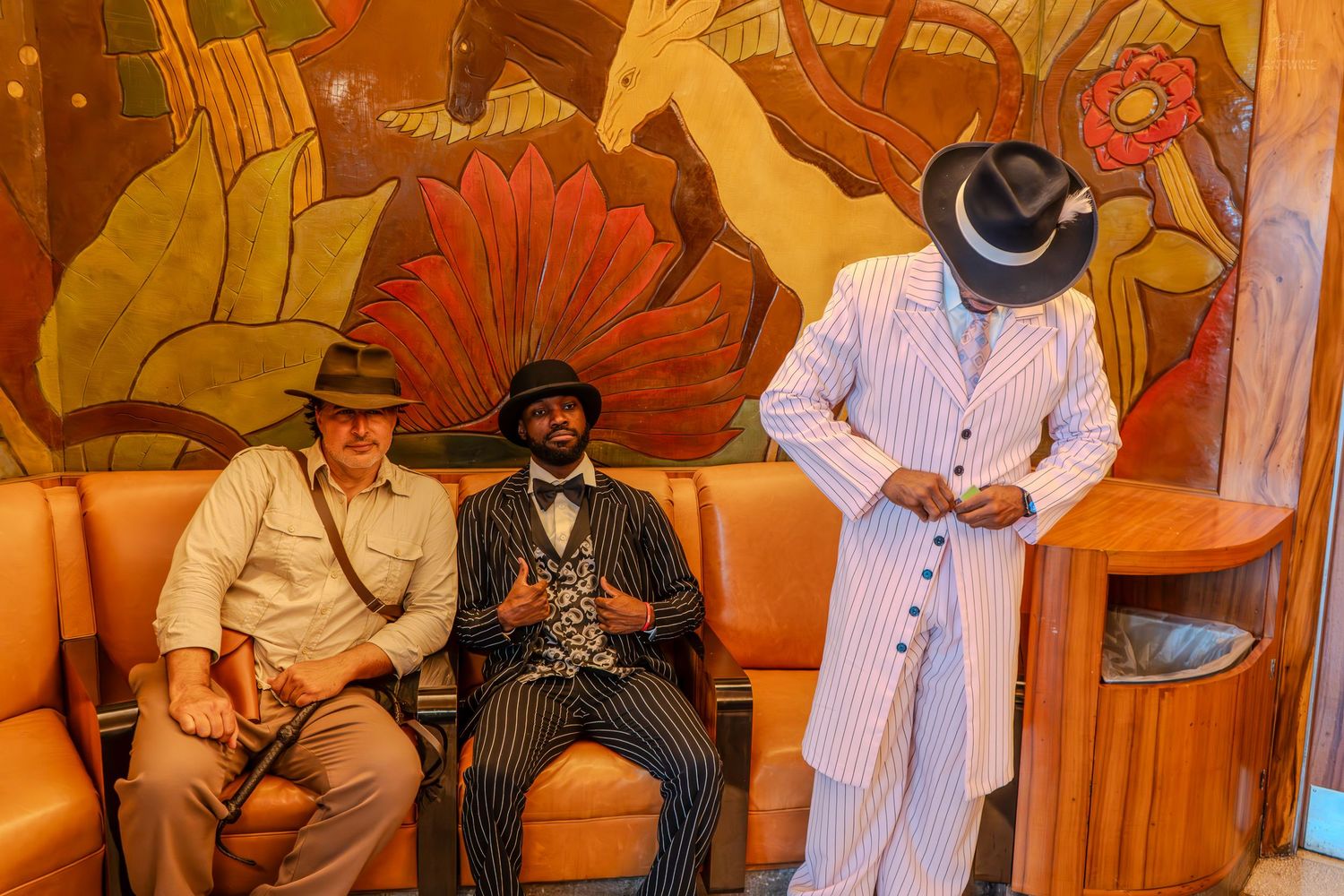 Three men in vintage zoot suits and fedora hats pose near a colorful mural in a retro-styled interior.