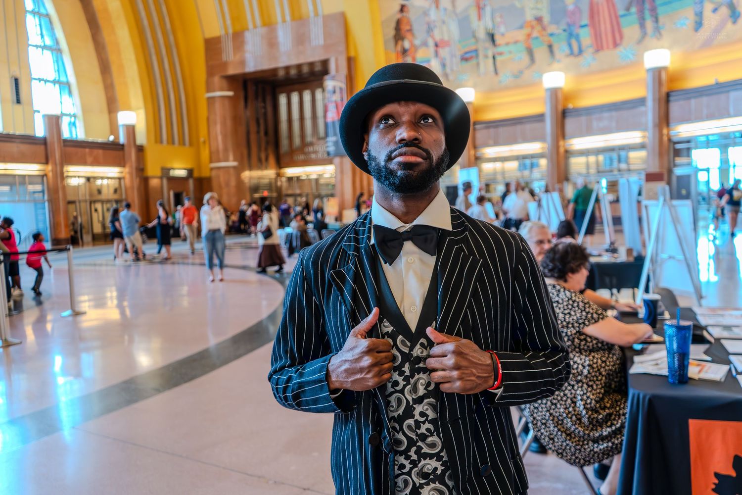 Man in vintage pinstripe suit and bowler hat poses in grand train station hall.