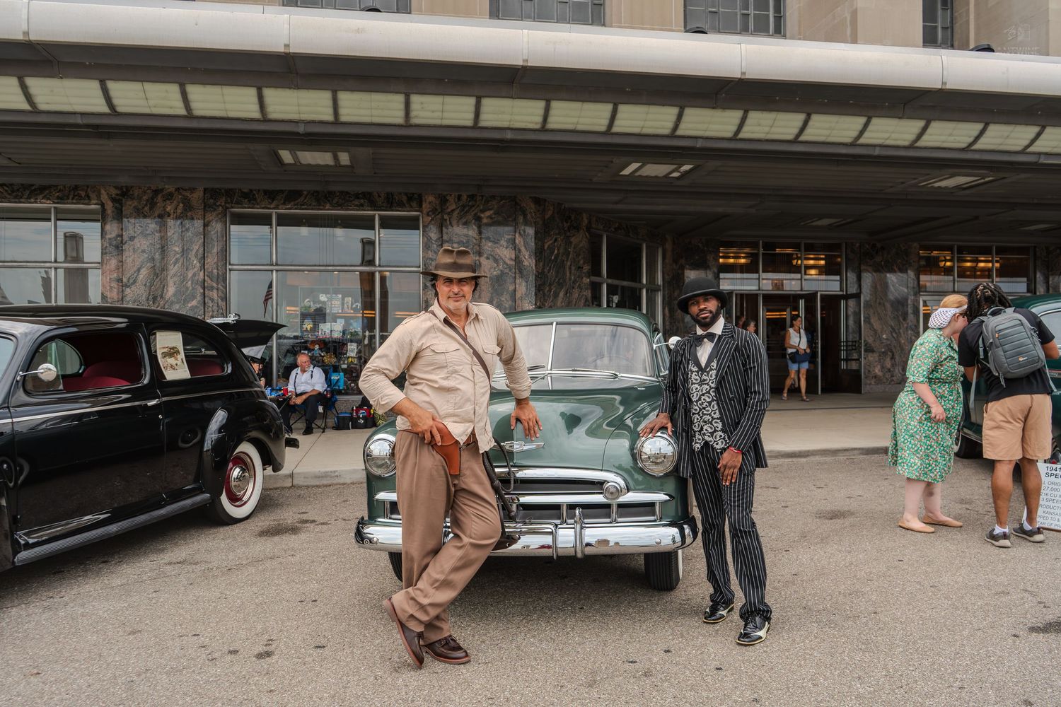 Two men in vintage 1940s attire pose beside a classic green car outside a retro building at a car show.