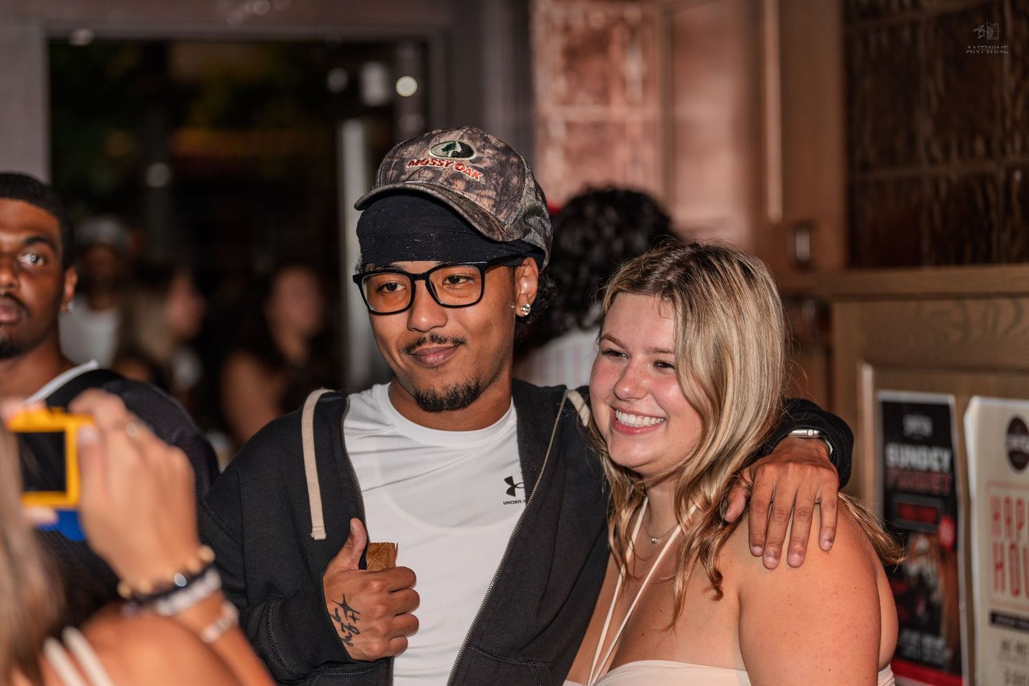 A man in a camo cap gives thumbs up while posing with a smiling blonde woman at a nightlife event.