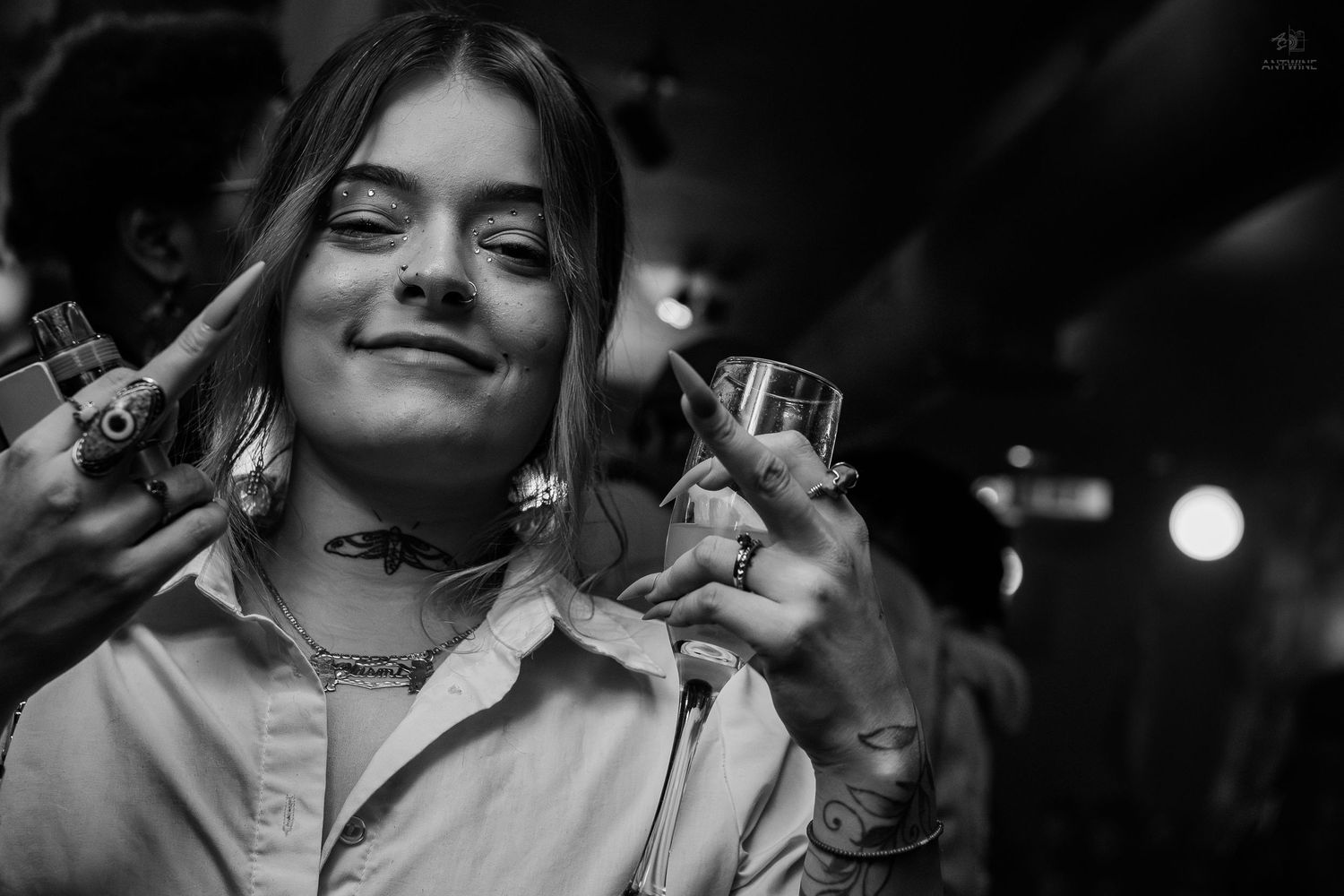 Black and white photo of a smiling young woman holding a shot glass at a party, flashing a peace sign.
