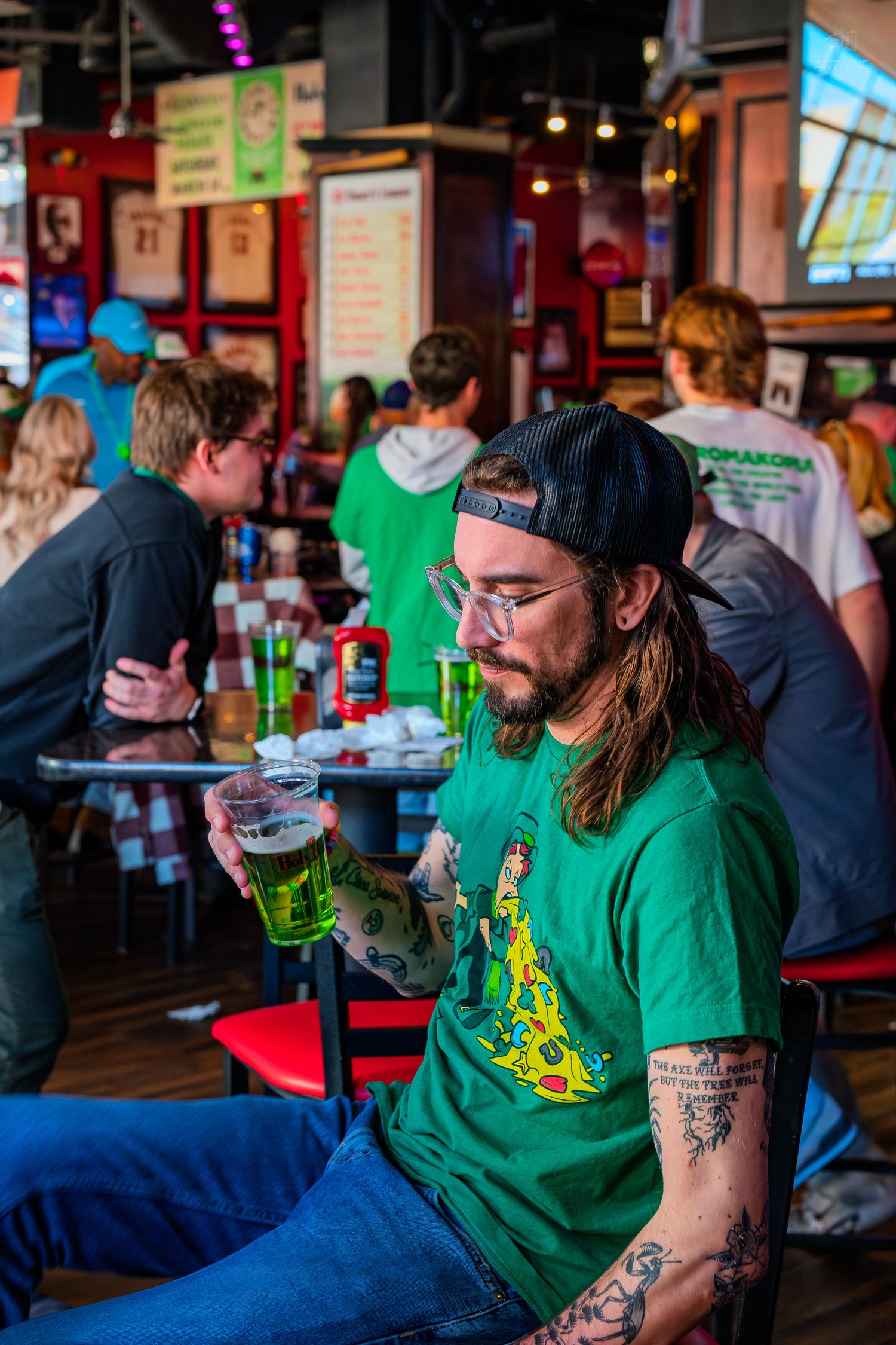 Tattooed man in green shirt holding green beer at a lively sports bar on St. Patrick's Day.