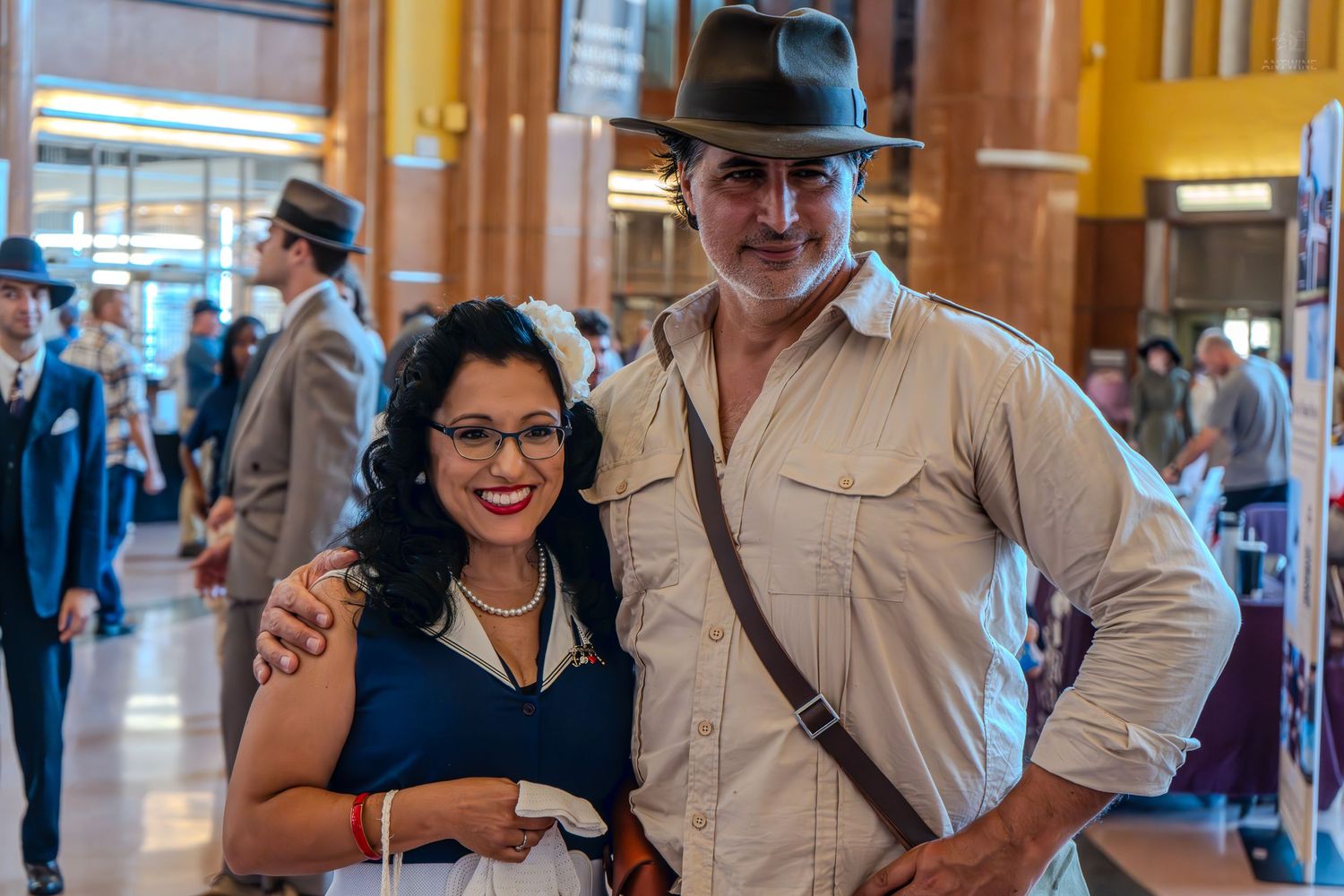 A couple in vintage 1940s costumes pose together at a retro-themed event inside an elegant venue.