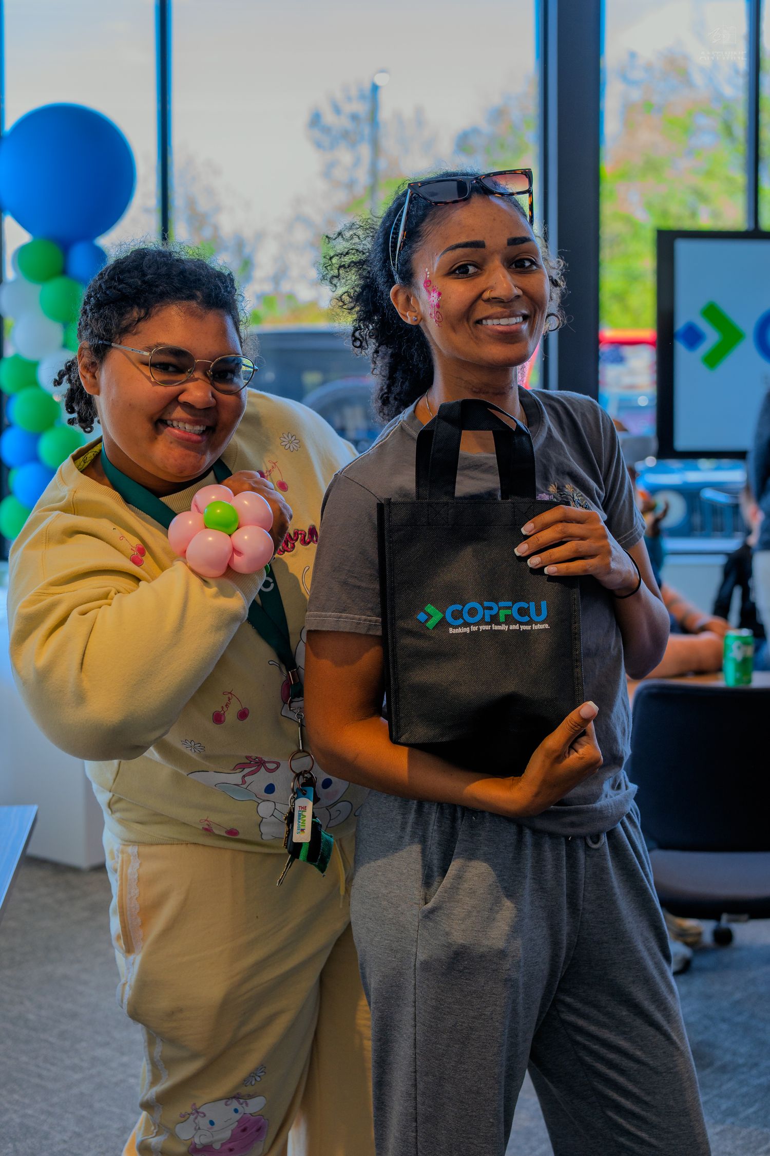 Two women smile holding a COPECU tote bag and balloon flower at a credit union promotional event.