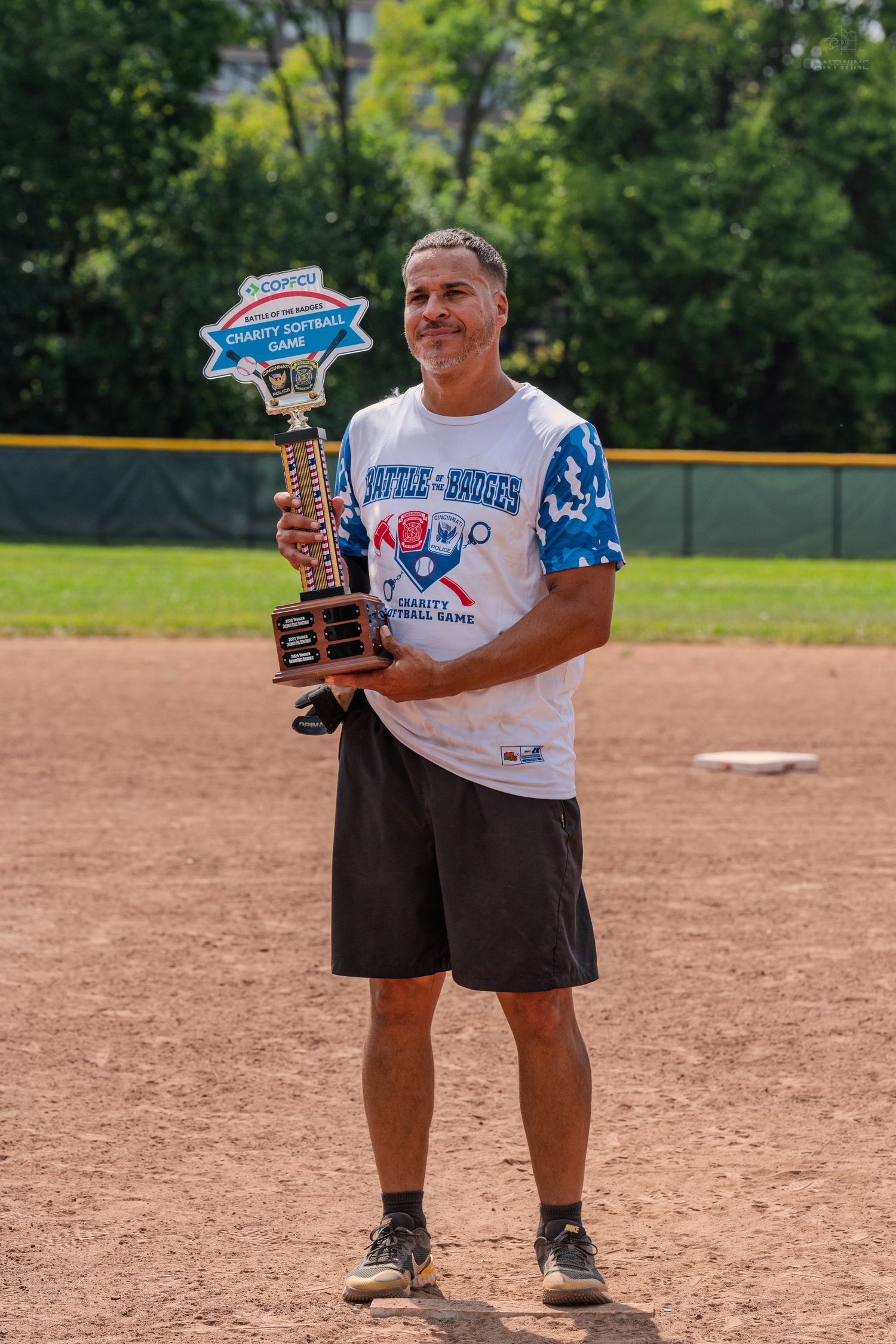 Smiling man poses with charity softball trophy on sunny baseball diamond in team uniform.