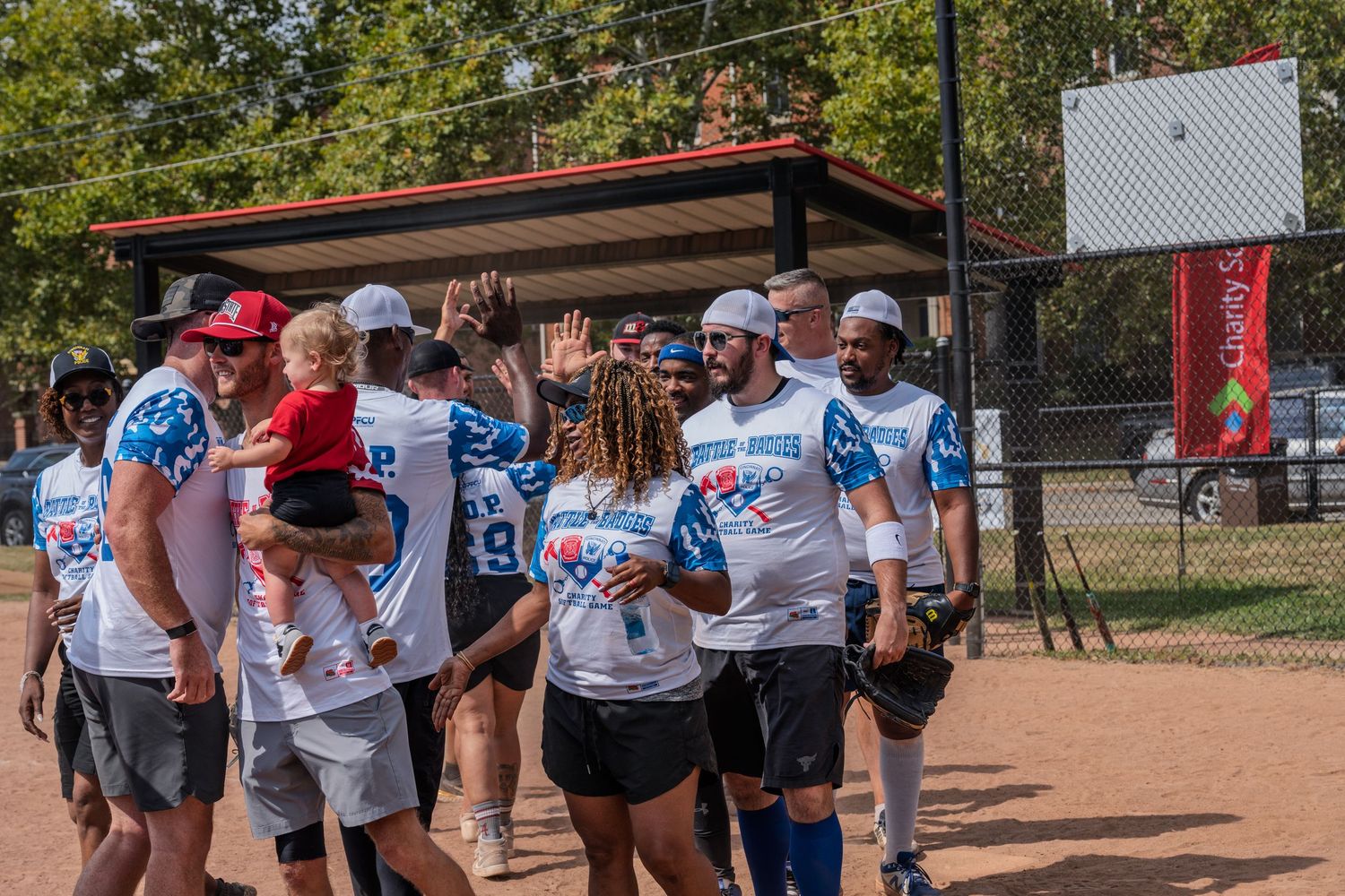 Softball team in white jerseys celebrating with high-fives on the field during a sunny outdoor charity game.