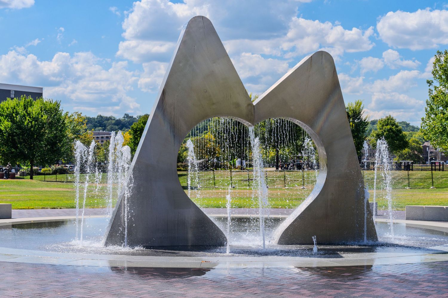 Modern concrete fountain sculpture with water jets in a sunny park setting with green trees and blue sky.