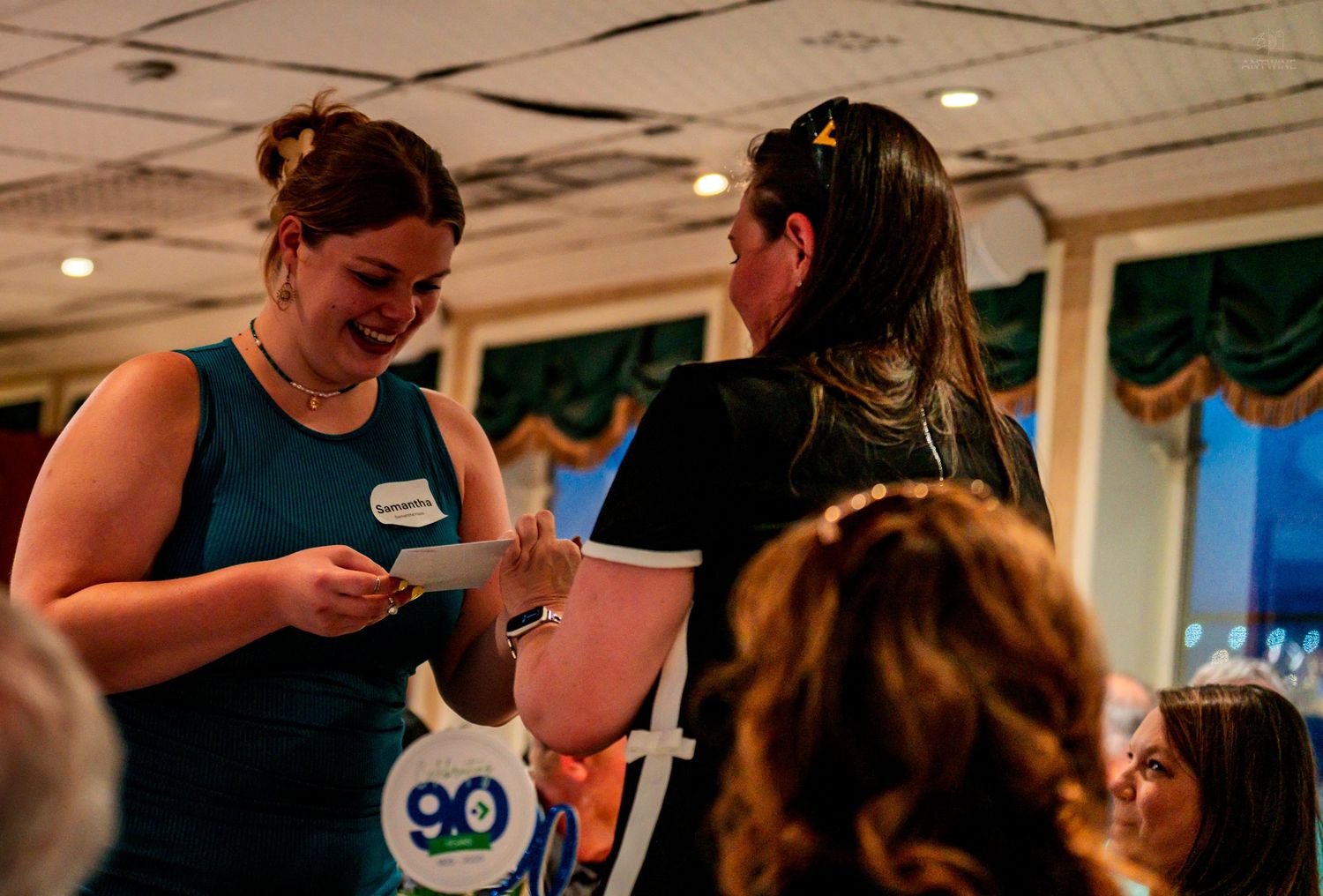 Two women exchanging a card and smiling at an indoor celebration event.