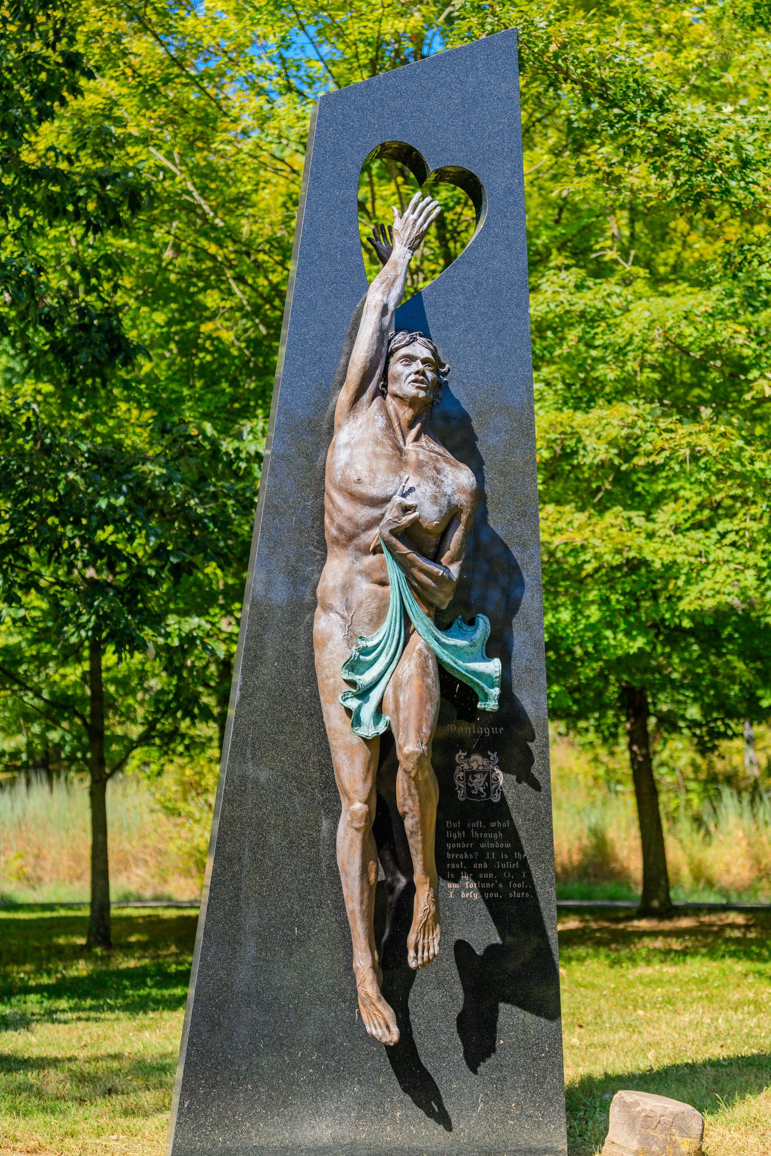 Bronze sculpture of a reaching male figure mounted on a dark granite obelisk with heart cutout, set in a lush green park.