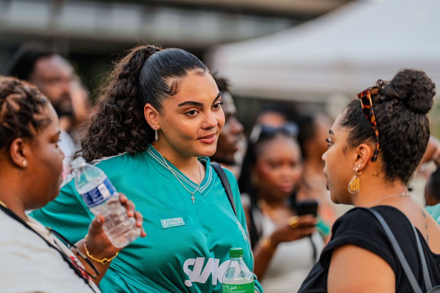 Young woman in a teal jersey holding water bottle chatting at a Juneteenth outdoor festival.