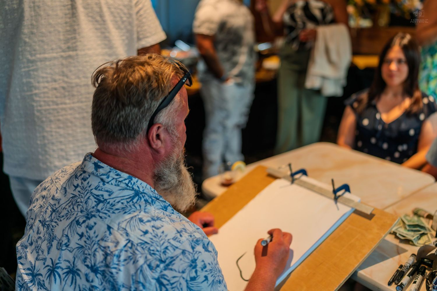 Man in floral shirt sketching at table while woman watches at indoor event.