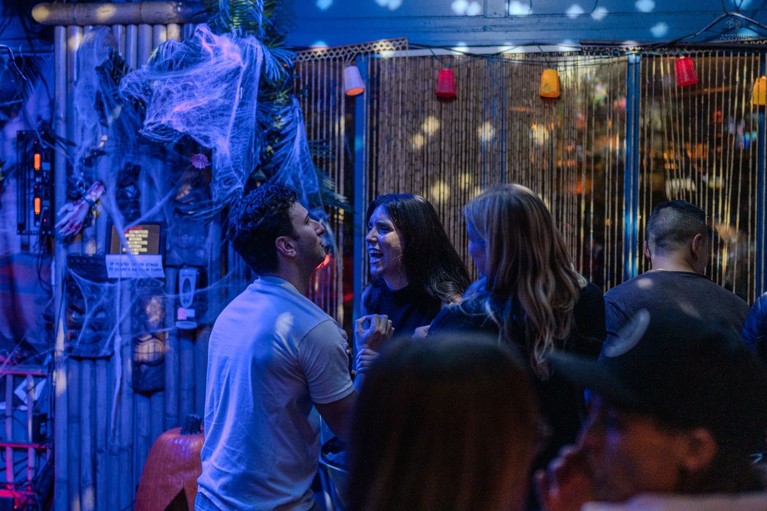 Group of people socializing under blue lights near Halloween decorations and string lights at a bar.