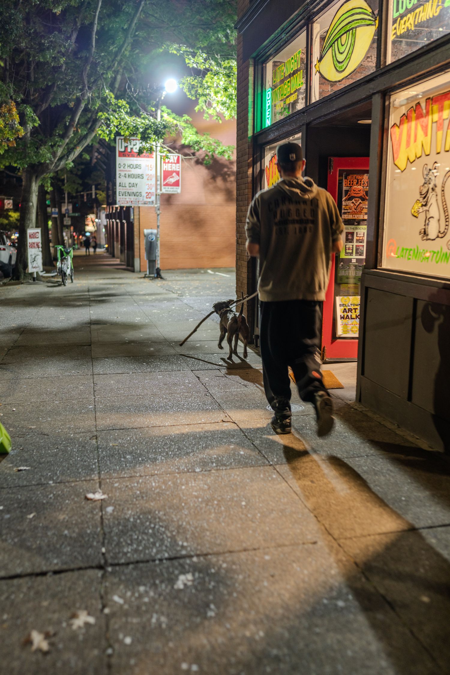 Man walking dog on leash past illuminated storefront on urban sidewalk at night.