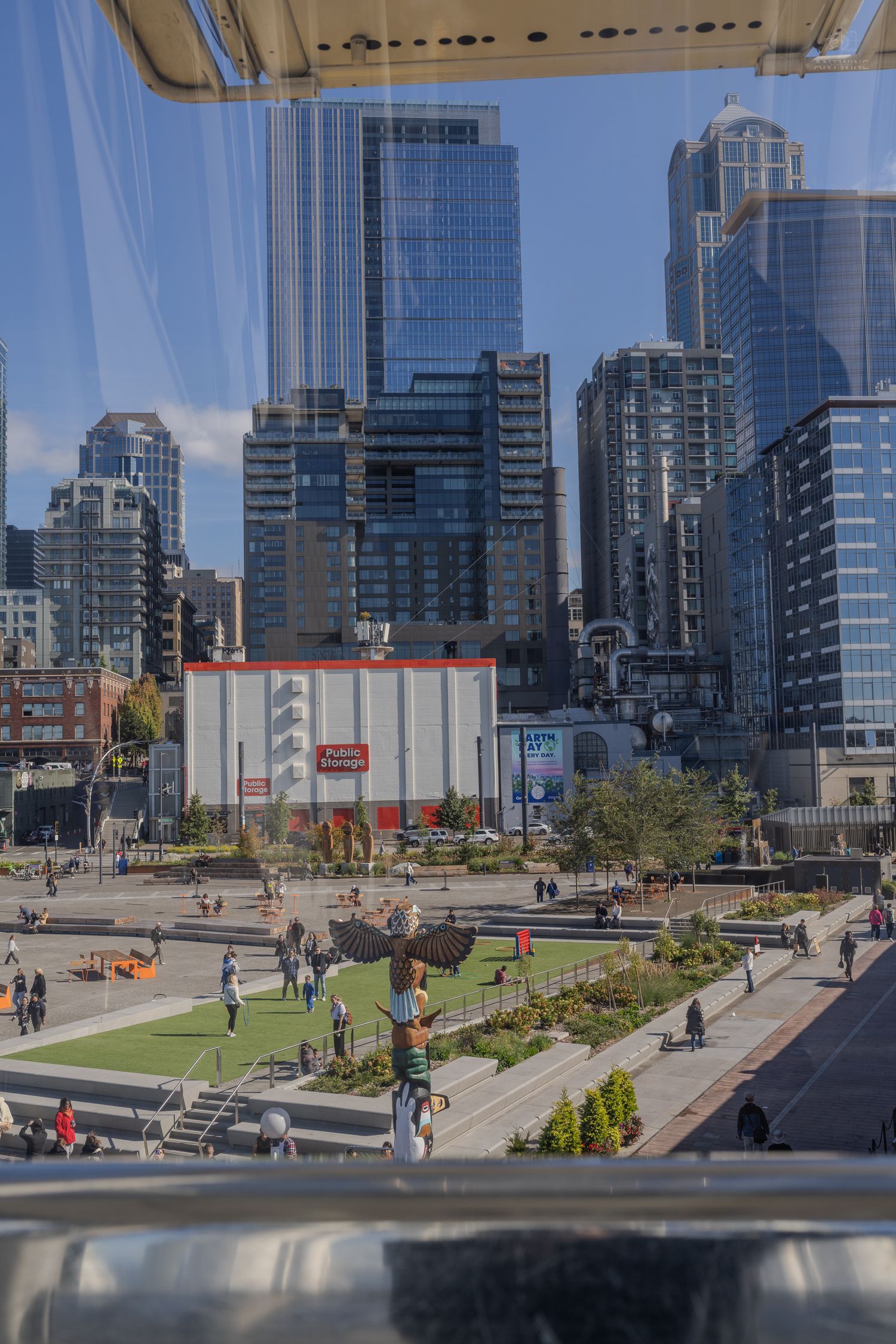 Aerial view through glass of Seattle's urban plaza with skyscrapers, green spaces, and pedestrians on sunny day.