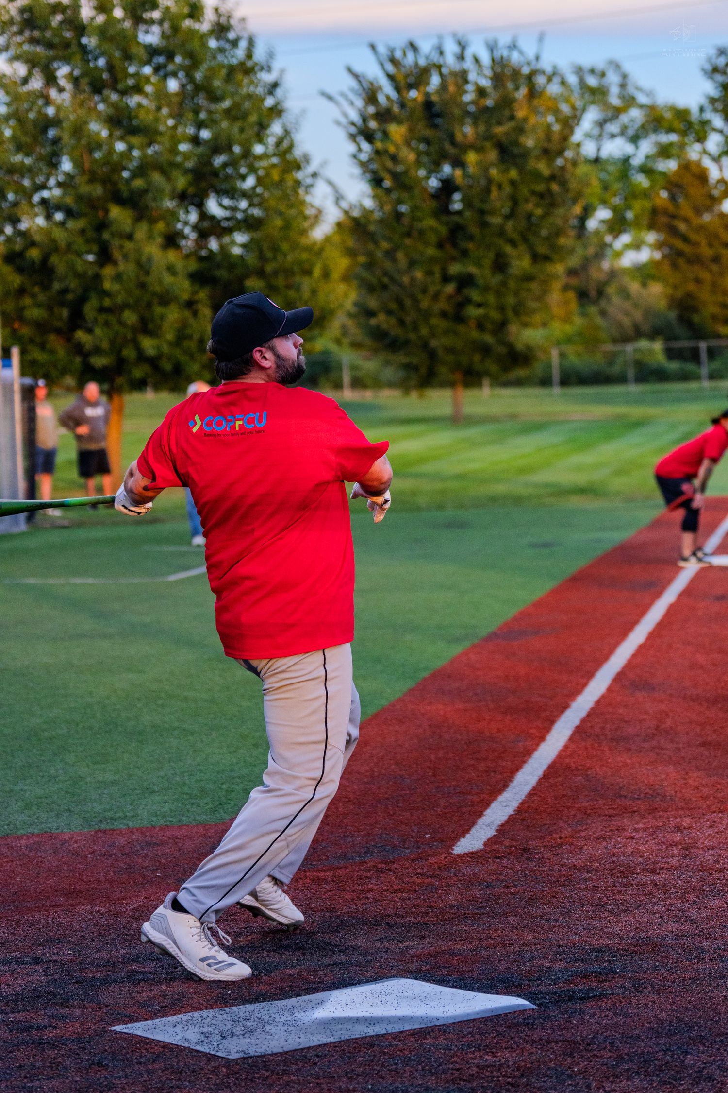 Man in red shirt running bases during softball game on red warning track at sunset.