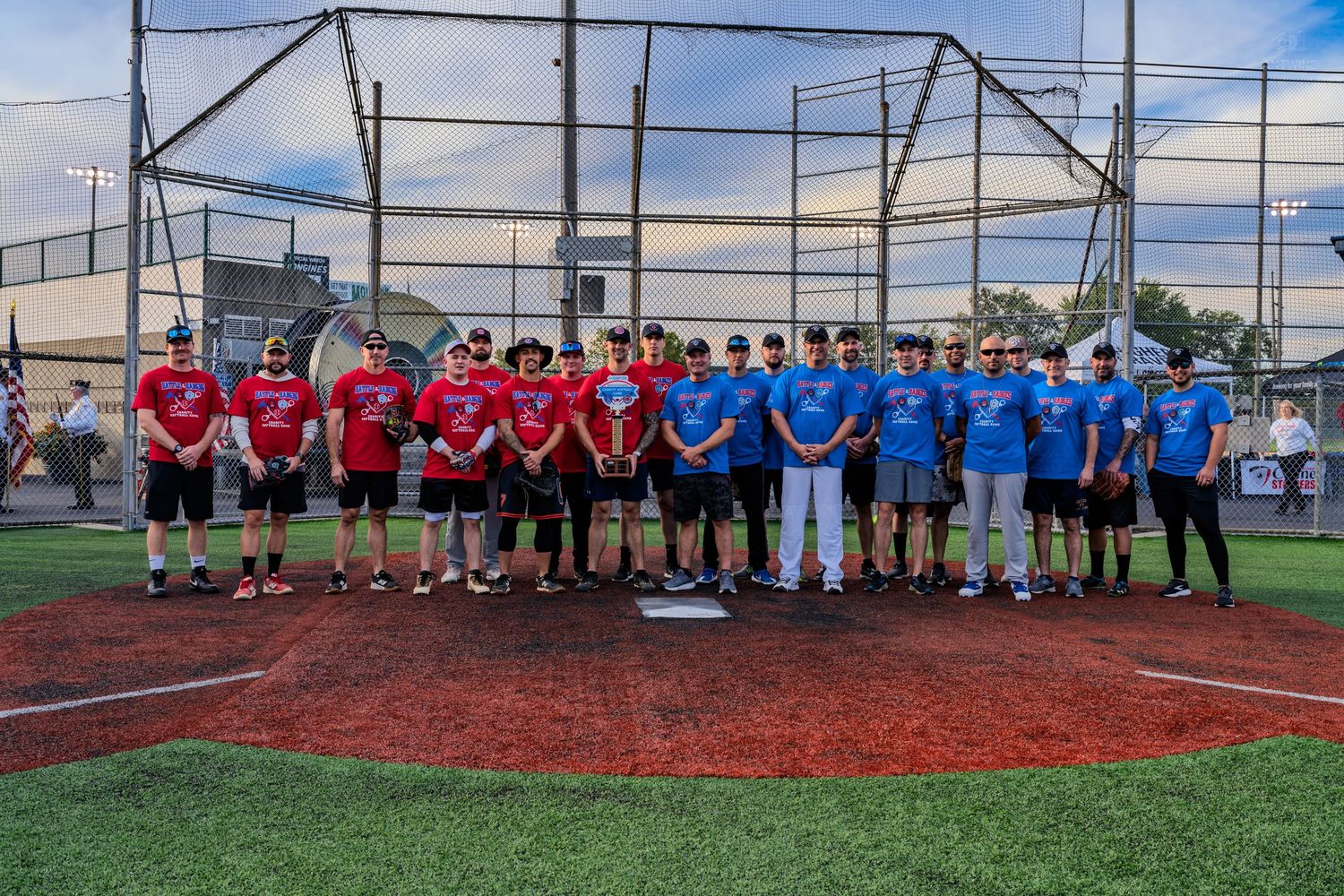 Two softball teams in red and blue shirts pose together on a baseball field at dusk.
