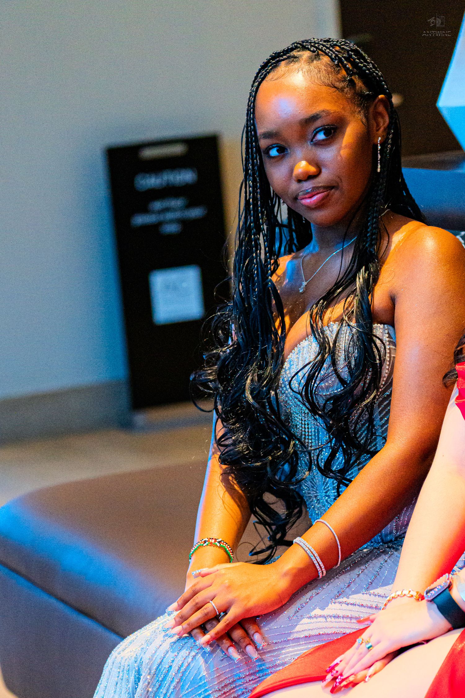 Young woman with long box braids in a silver sequin dress seated at a formal event.