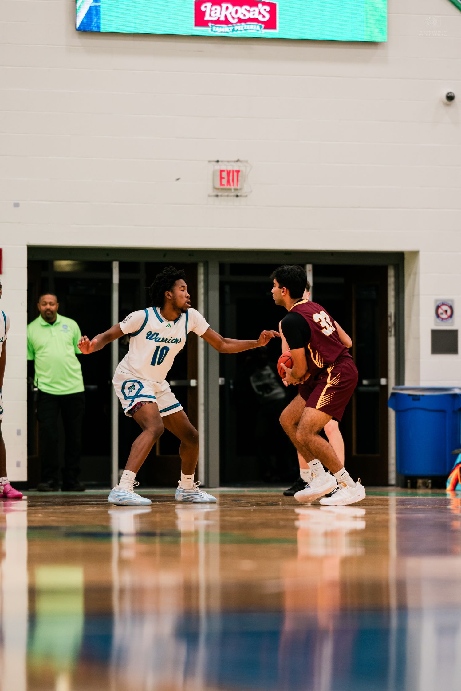 Warriors player number 10 defends against a maroon-jersey opponent dribbling near the gym entrance.