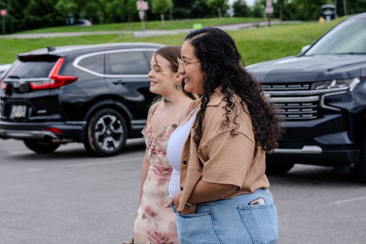 Two women smile and walk together through a parking lot, one in a floral dress and one in a tan top.