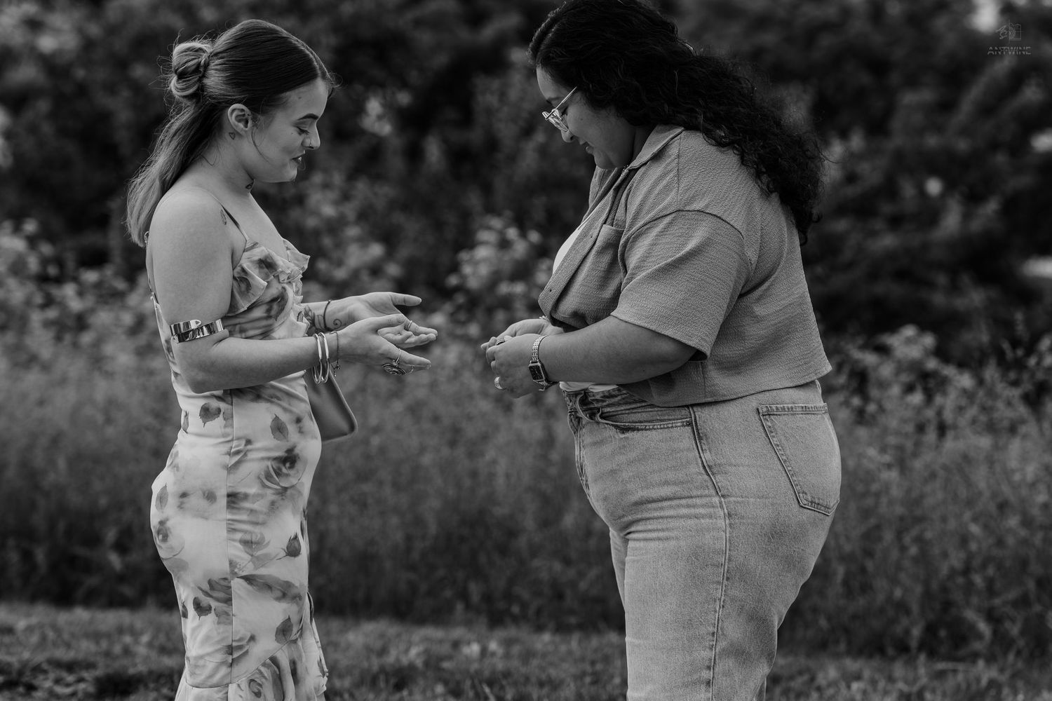Two women exchange something outdoors in a garden setting, captured in black and white photography.
