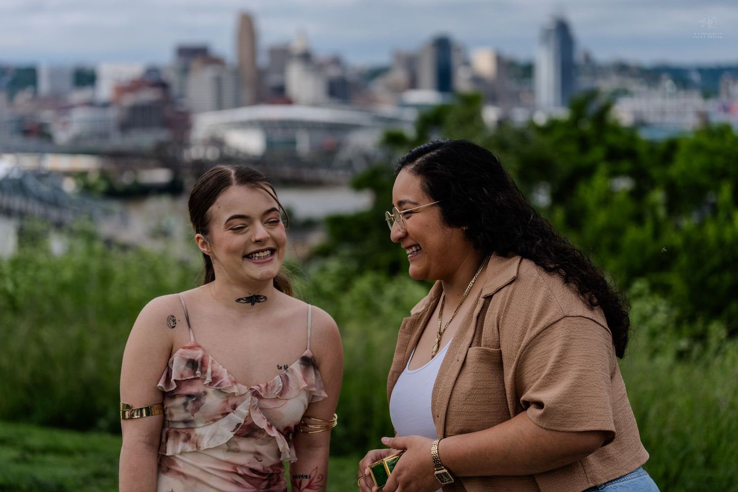 Two women smiling and laughing outdoors with a city skyline visible in the background.