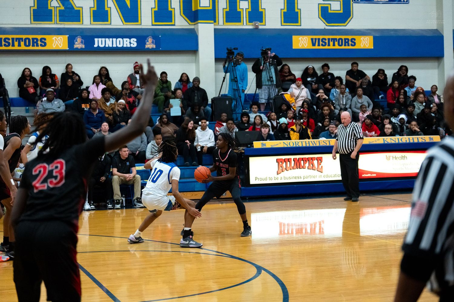 Blue jersey player number 10 dribbles against red jersey defender during packed high school basketball game.
