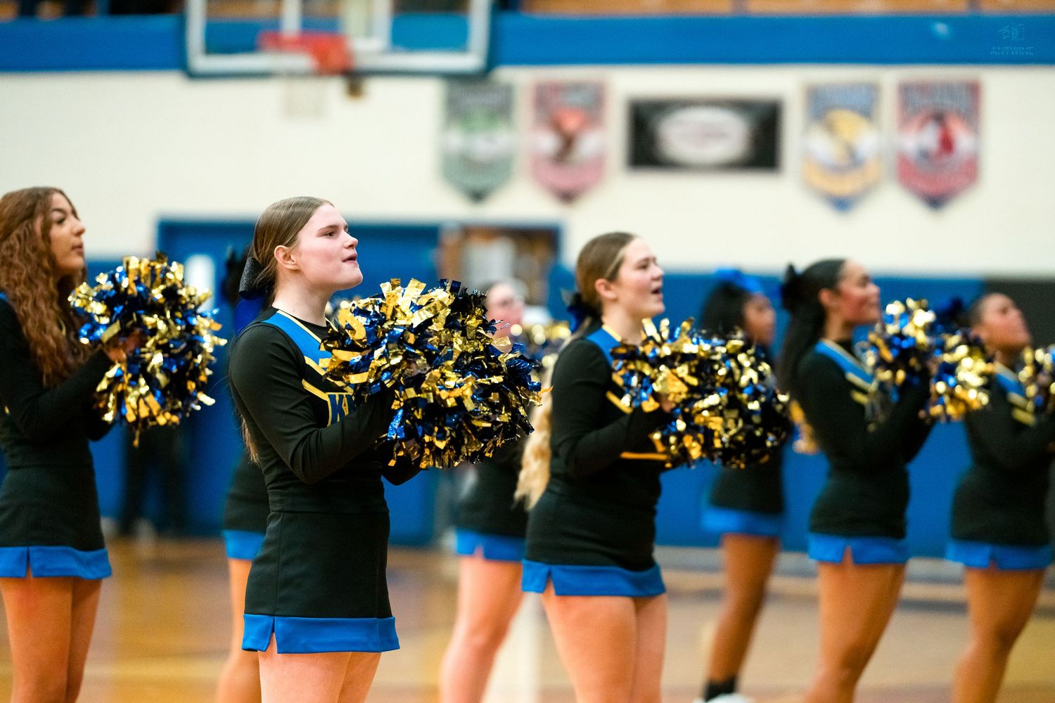 High school cheerleaders in black and gold uniforms perform with pom-poms during a basketball game.