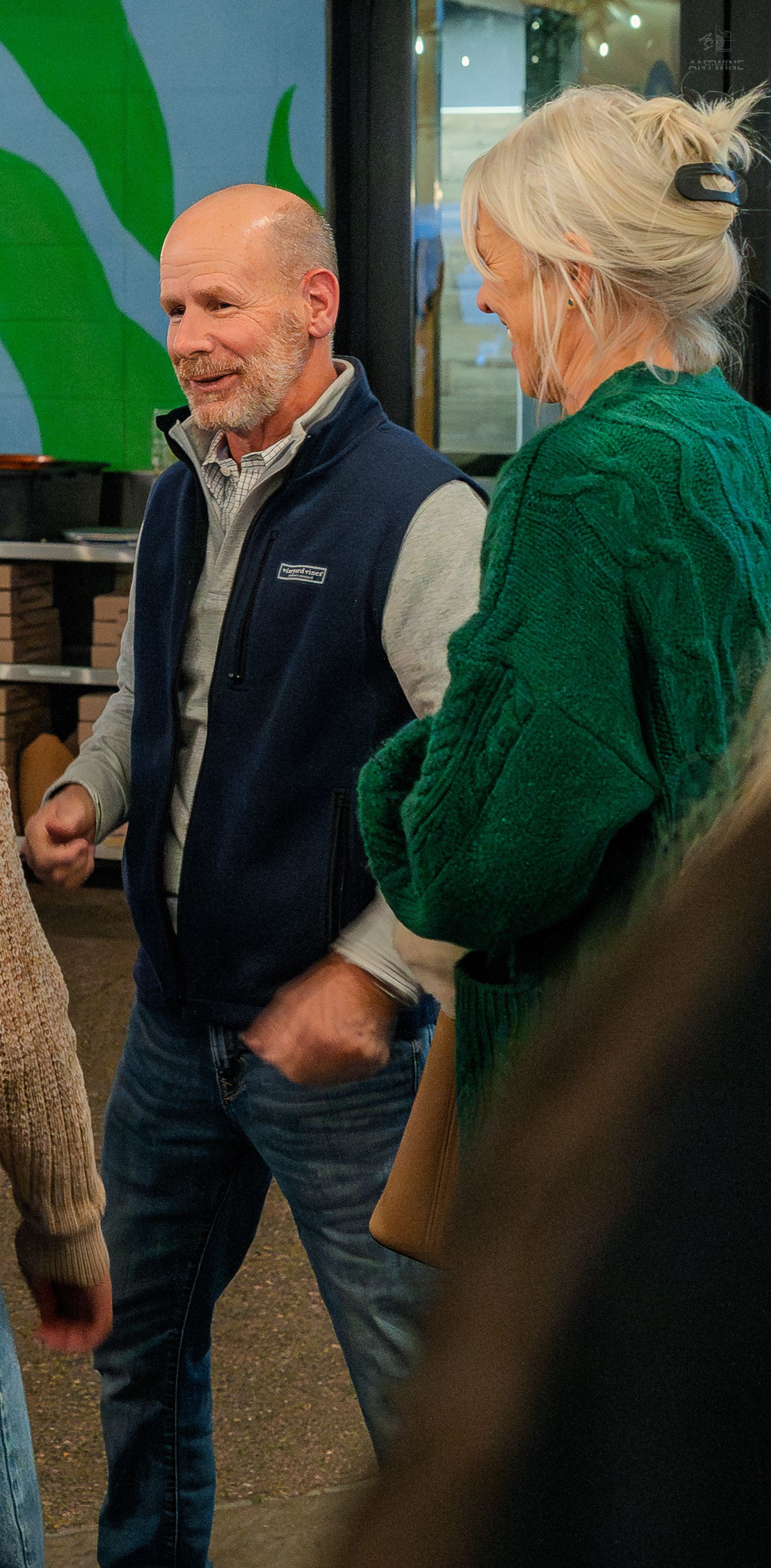 Older man in navy vest and blonde woman in green sweater chatting at an indoor event.