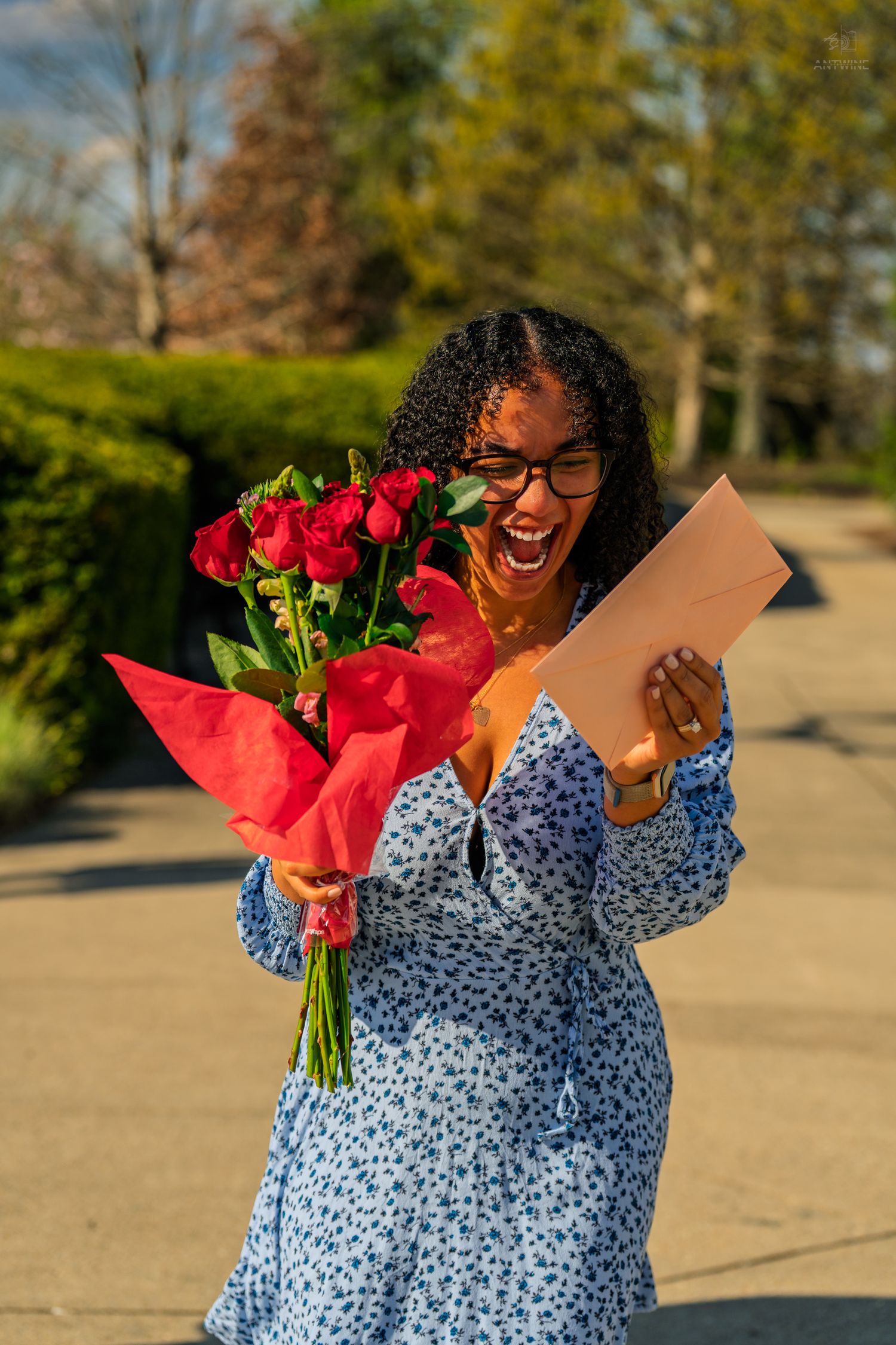 A delighted woman in a blue floral dress reads a letter while holding a bouquet of red roses outdoors.