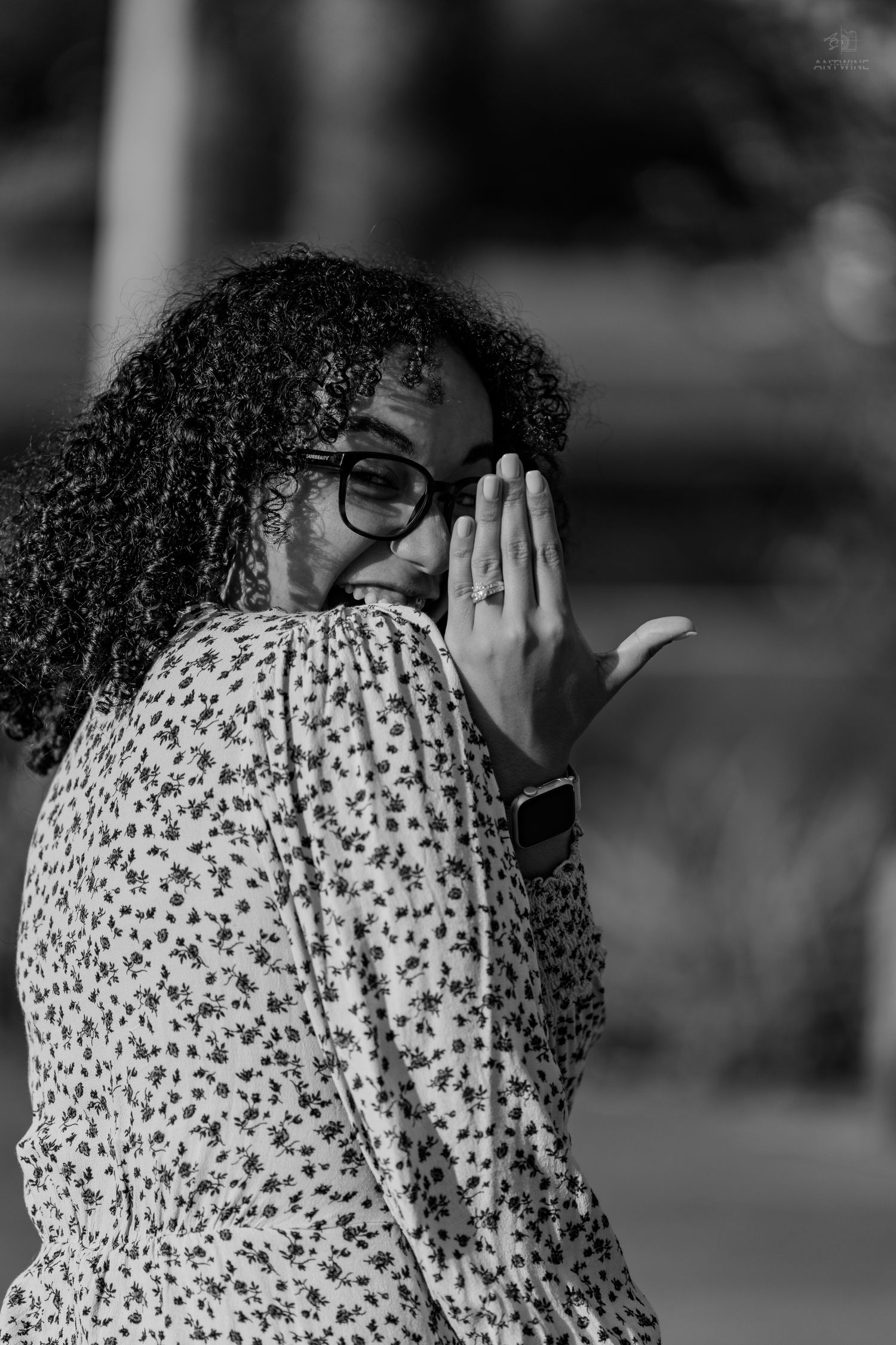 Black and white portrait of a woman with curly hair and glasses, laughing while covering her mouth.