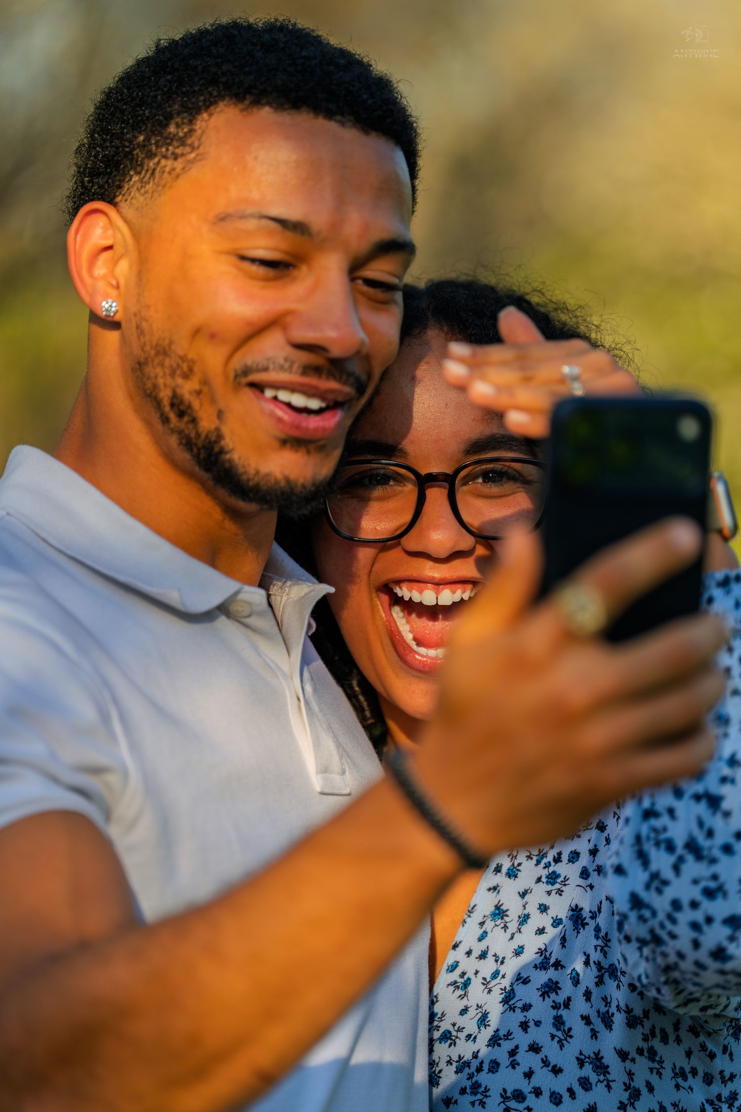 Laughing engaged couple taking a selfie in sunny park, woman showing engagement ring while man embraces her.