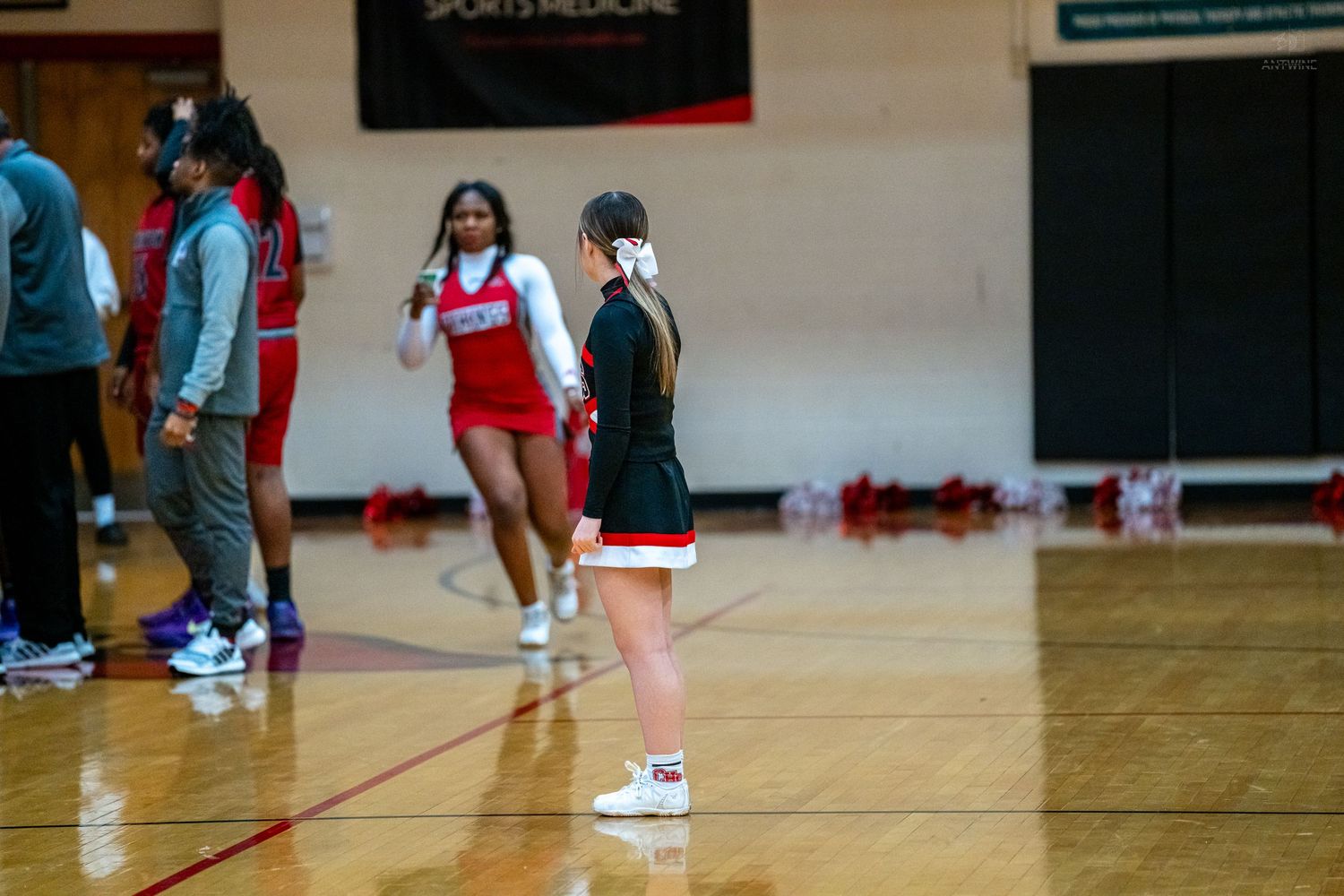 A cheerleader in black and white uniform stands on the basketball court sideline during a game.