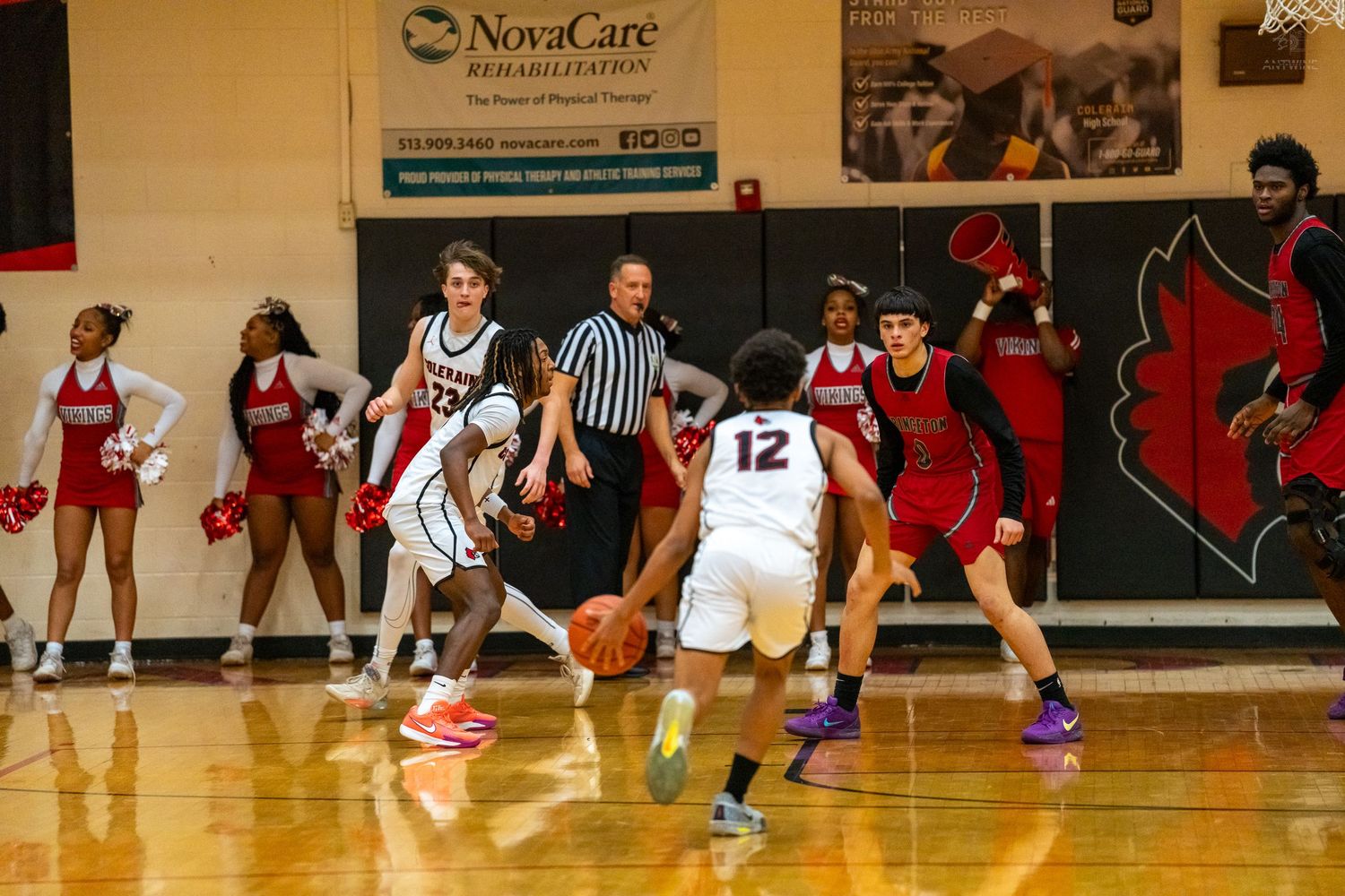 Basketball players compete near cheerleaders as referee watches indoor game.