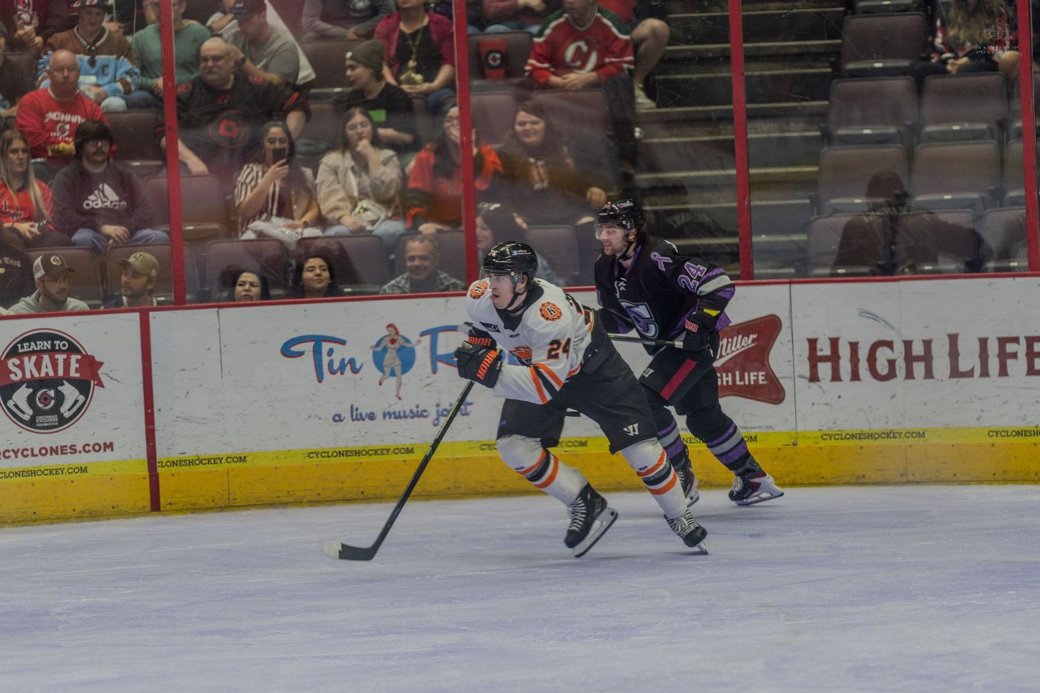 Hockey player #24 in orange uniform battles dark-jersied defender for puck possession during indoor ice hockey game.