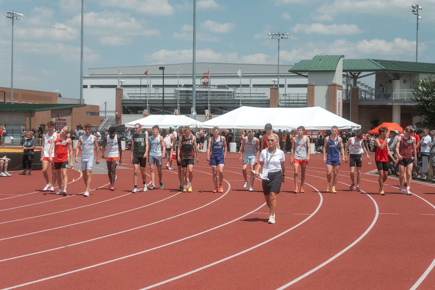 Track and field competitors walk on a red track at an outdoor stadium during a sunny day event.