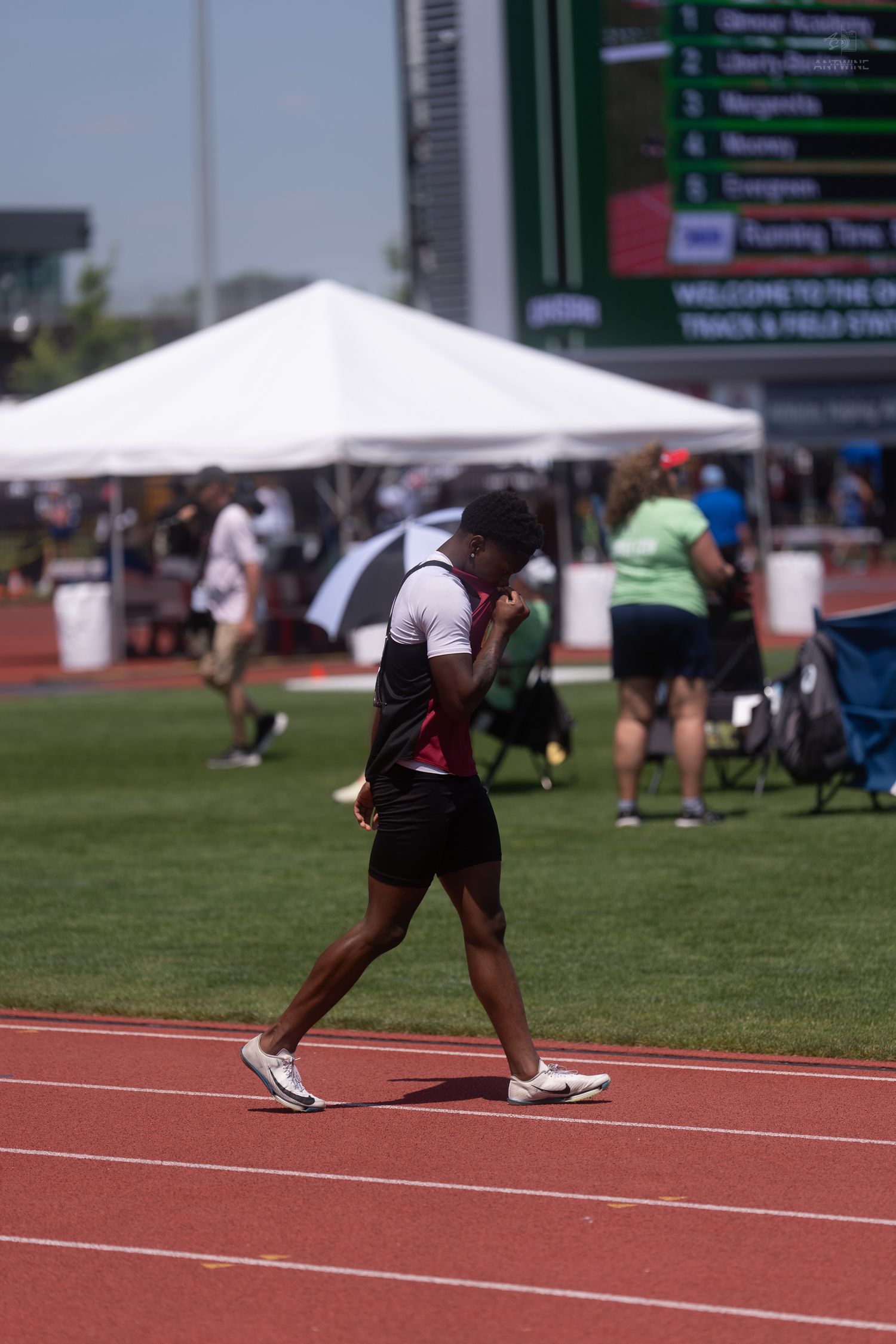 Female athlete in black uniform walks on a red track near a scoreboard at an outdoor track and field championship meet.