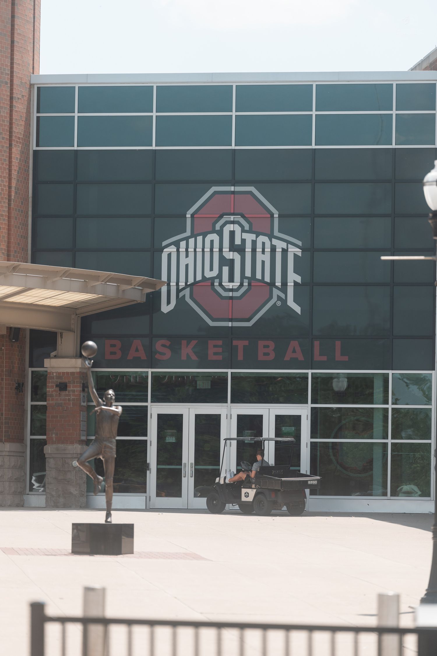 Ohio State Basketball arena entrance featuring bronze player statue and large Buckeyes logo on glass facade.