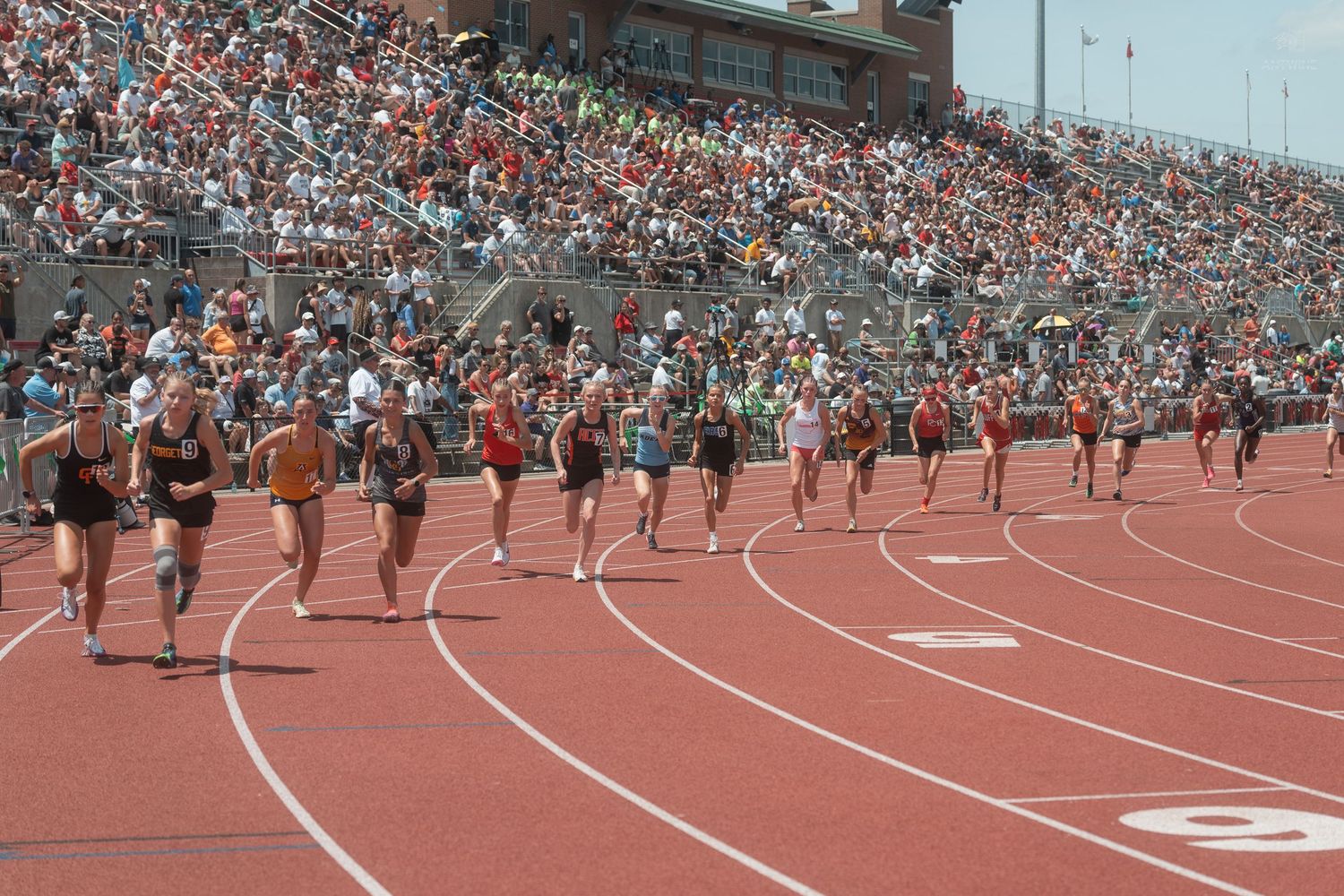 Female runners sprint down the track during a competitive high school track and field meet with a full stadium.