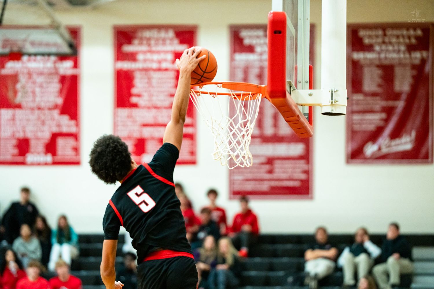 Player #5 in black uniform reaches up to complete slam dunk during basketball game.