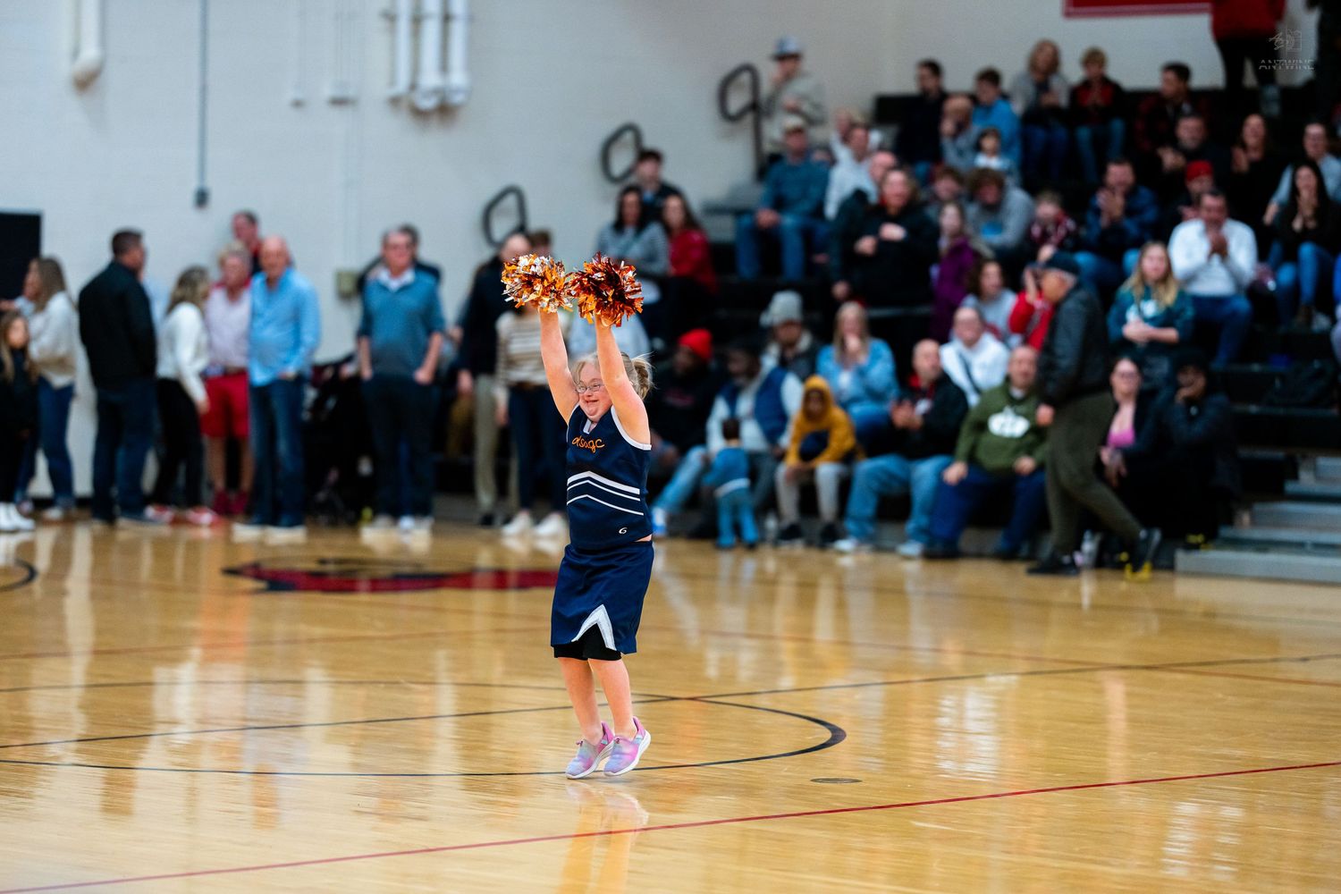 Lone cheerleader in navy uniform performs with gold pom-poms on basketball court before packed stands.