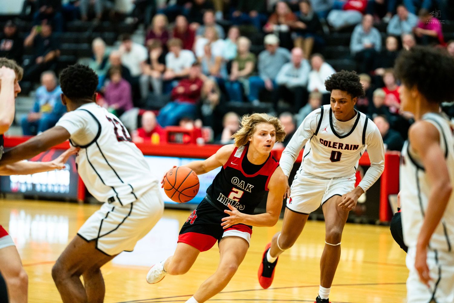 High school basketball player number 2 in white drives between two defenders during a competitive indoor game.