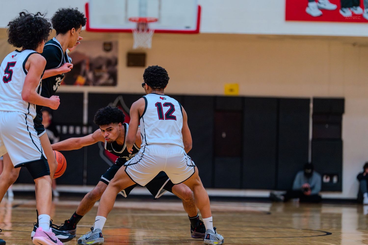 High school basketball players compete for the ball as player #12 defends against a driving opponent in a gym.
