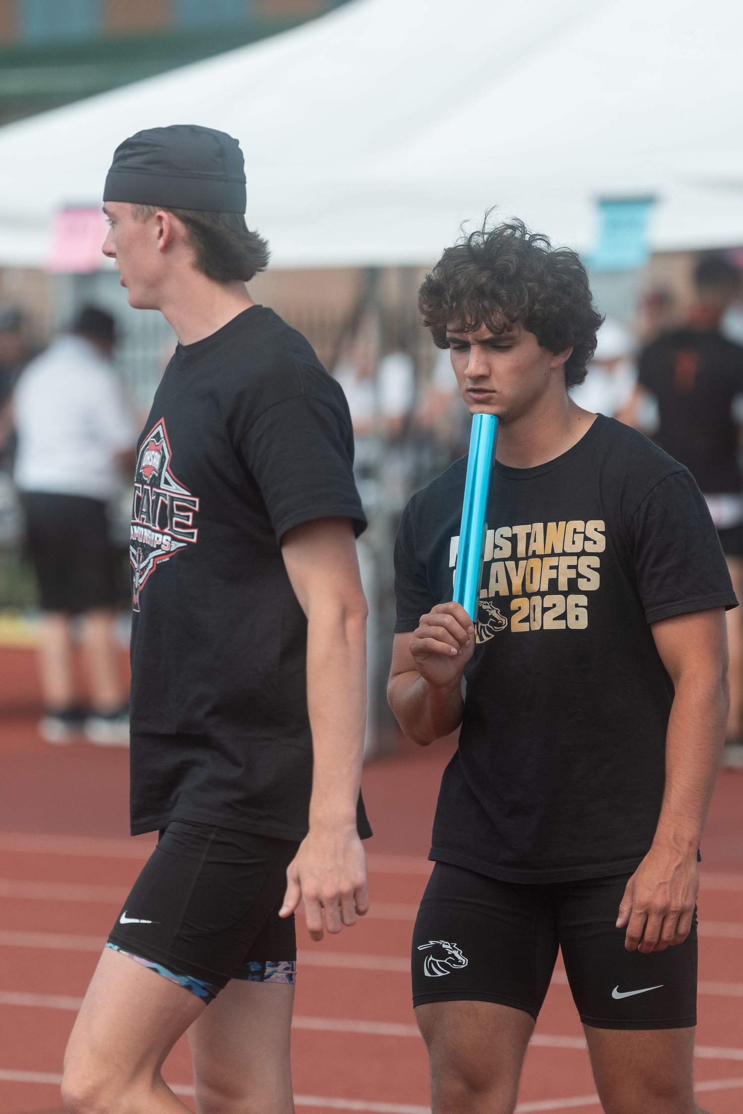 Two young male relay runners in black Mustangs Playoffs shirts prepare on a red track, one holding a blue baton.
