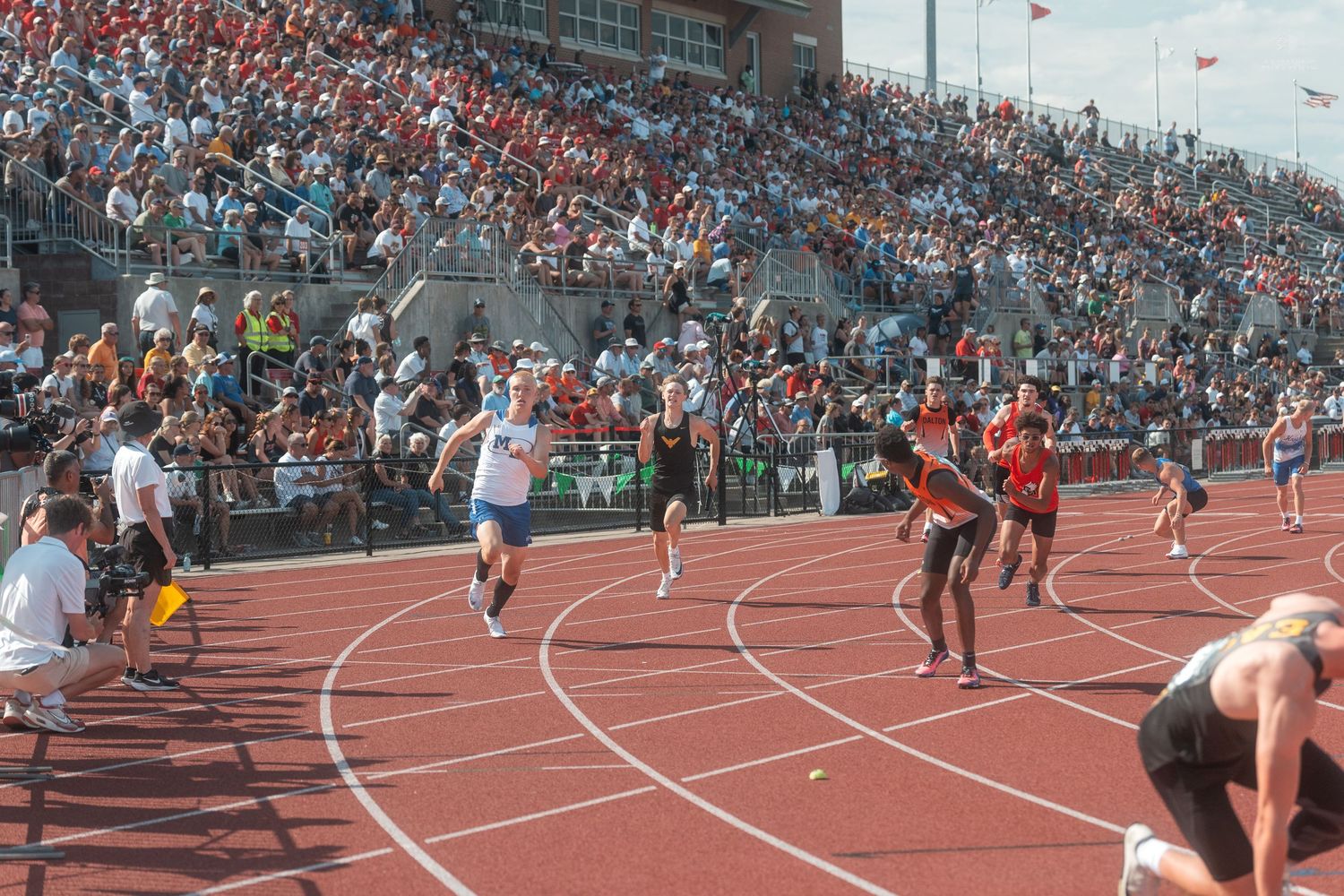 Runners sprint down a red track during a relay race at a crowded outdoor high school track and field meet.