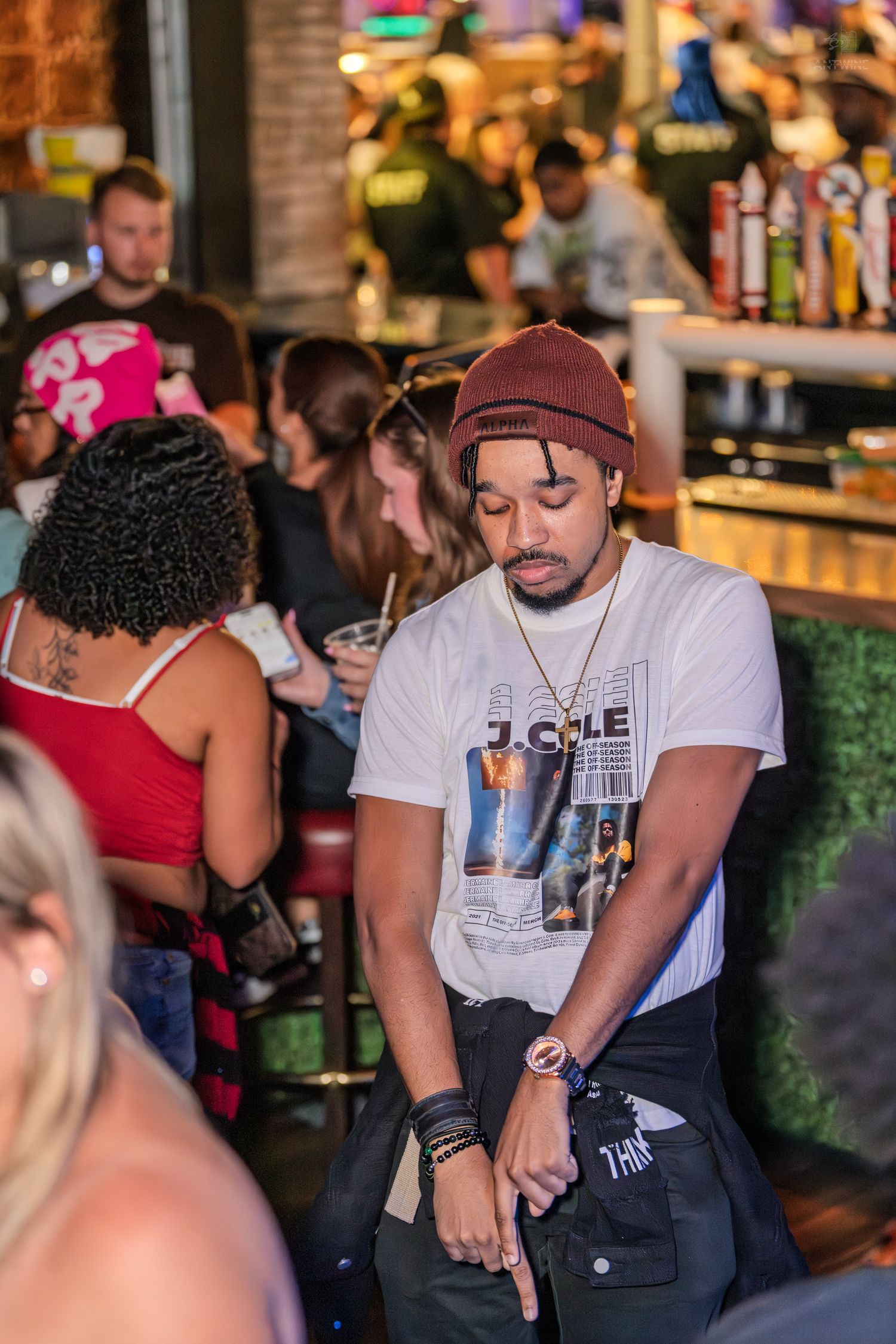 Young man in J. Cole shirt and burgundy beanie relaxing at a busy bar.