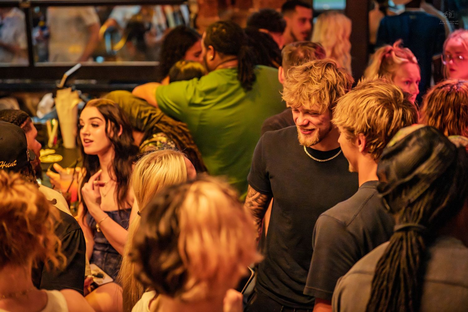 Man and woman in close conversation at a warm, golden-lit crowded bar or social venue.