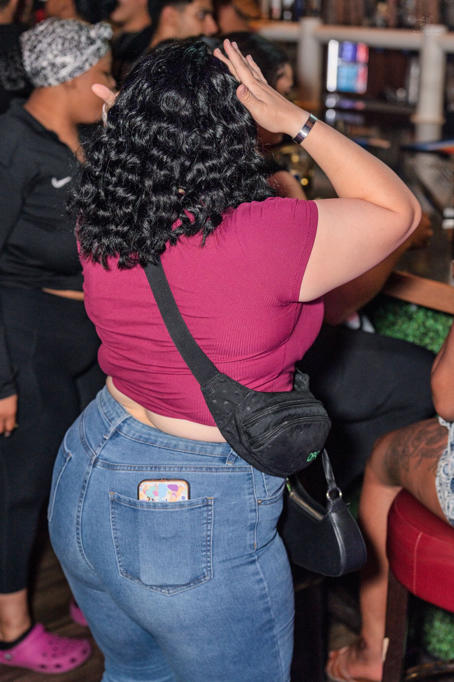 Curly-haired woman in pink crop top and jeans with crossbody bag at a nightclub.