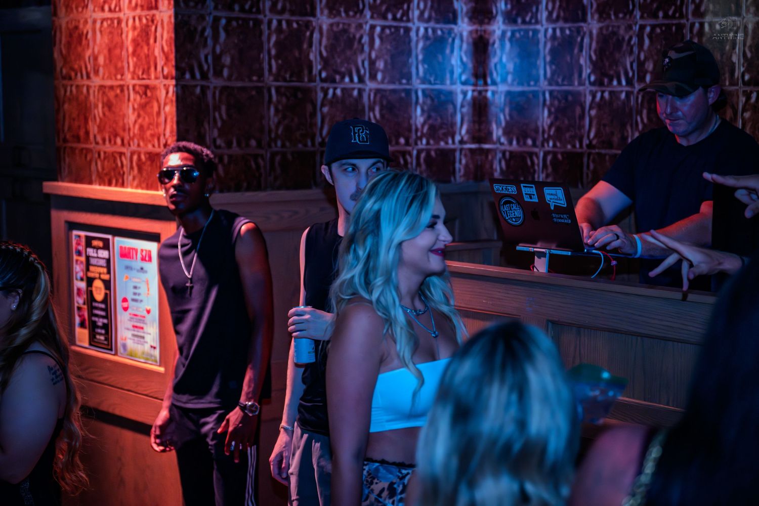 People gather near a DJ booth under colorful neon lights at a nightclub.