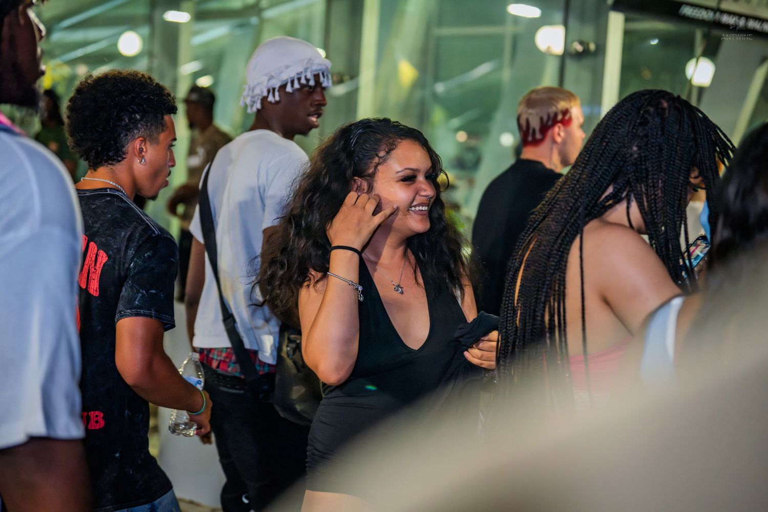 A smiling woman with curly hair in a black top enjoys a lively indoor event with a diverse crowd.